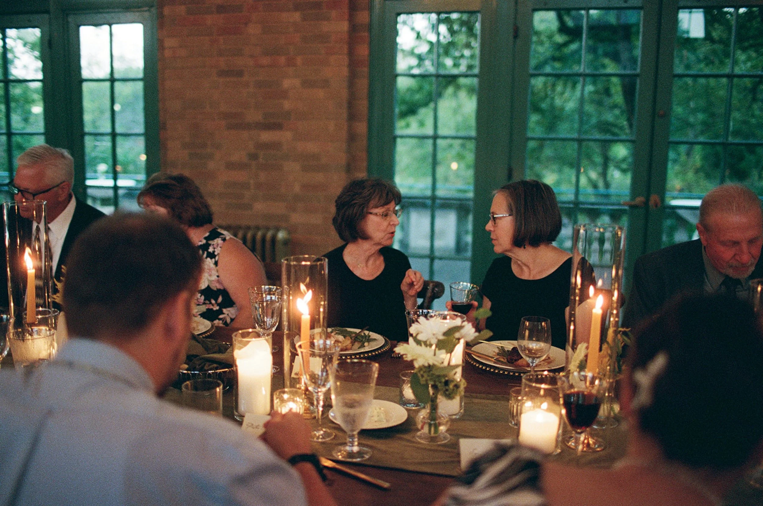 Guests lean across the candlelit table in easy conversation — the warm brick walls and teal window frames of Columbus Park Refectory creating an intimate dinner-party atmosphere