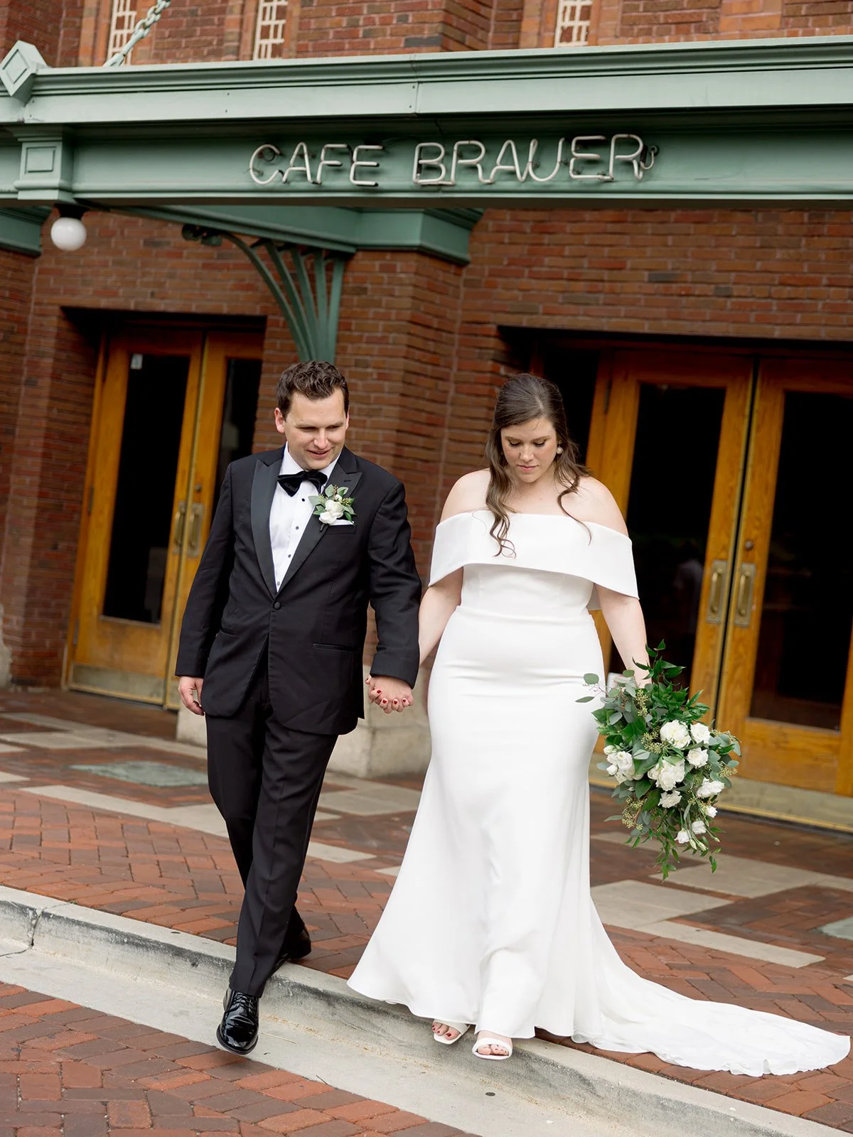 Couple walking hand-in-hand along the brick courtyard in front of Cafe Brauer, the green-trimmed entrance above them.
