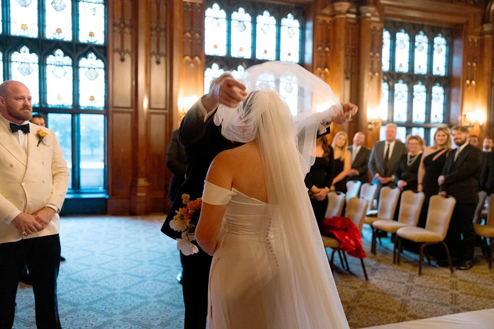 A loved one lifts the bride's veil in a tender, almost ceremonial gesture inside the University Club Chicago chapel — stained glass windows glowing behind them, the groom watching nearby.