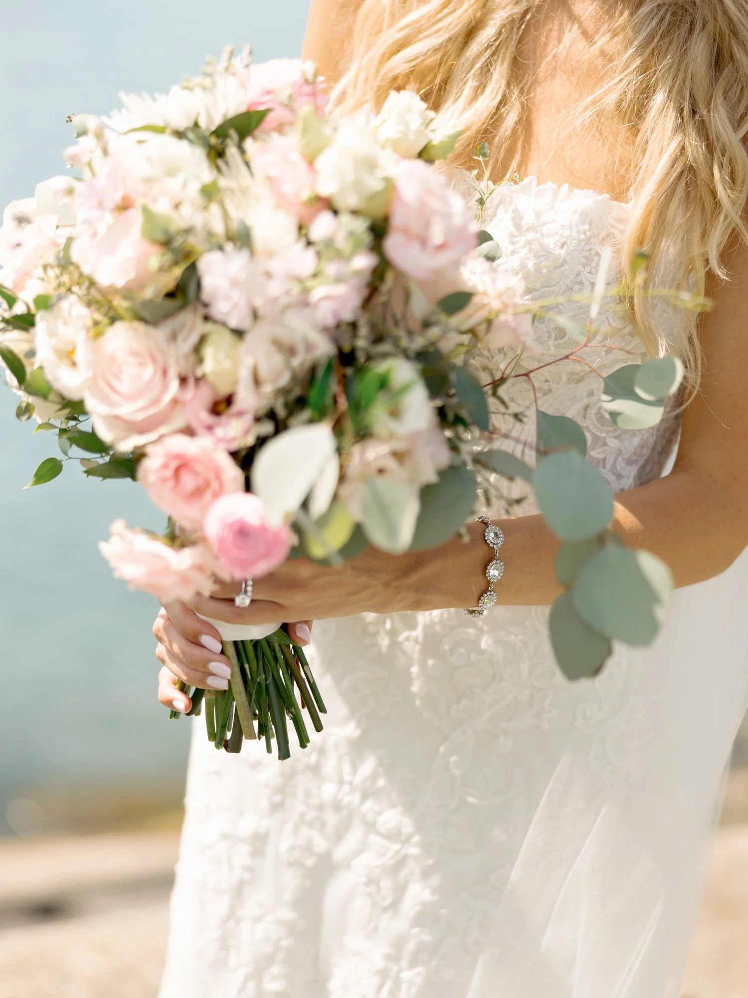 A lush bridal bouquet of blush roses, ranunculus, and eucalyptus held close by the bride at Olive Park, her diamond bracelet glinting in the sunlight.