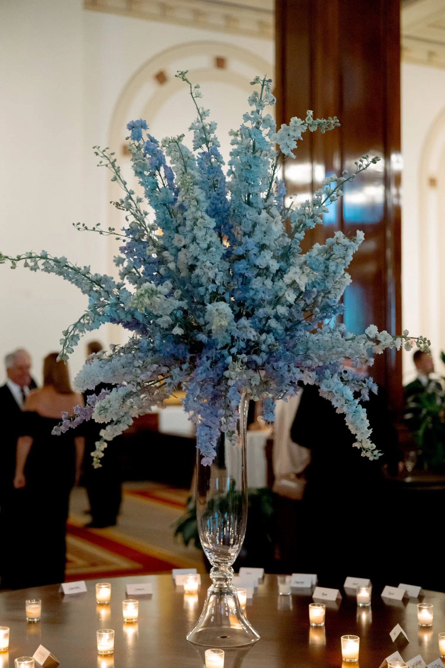 A dramatic tower of blue delphiniums anchors the escort card table, surrounded by rows of glowing votive candles at the Union League Club of Chicago.
