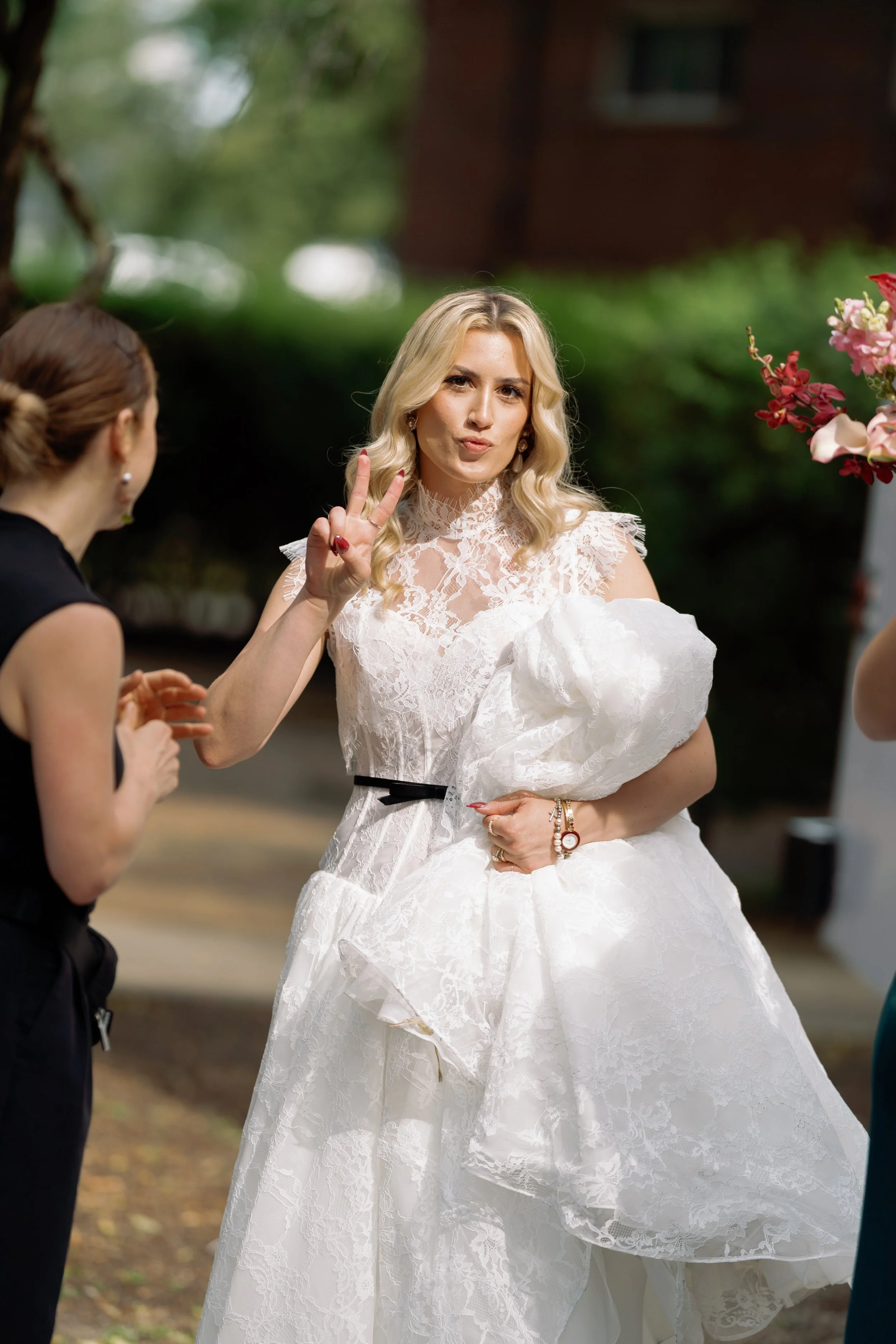 Bride flashes a peace sign outdoors while holding her red and blue bouquet