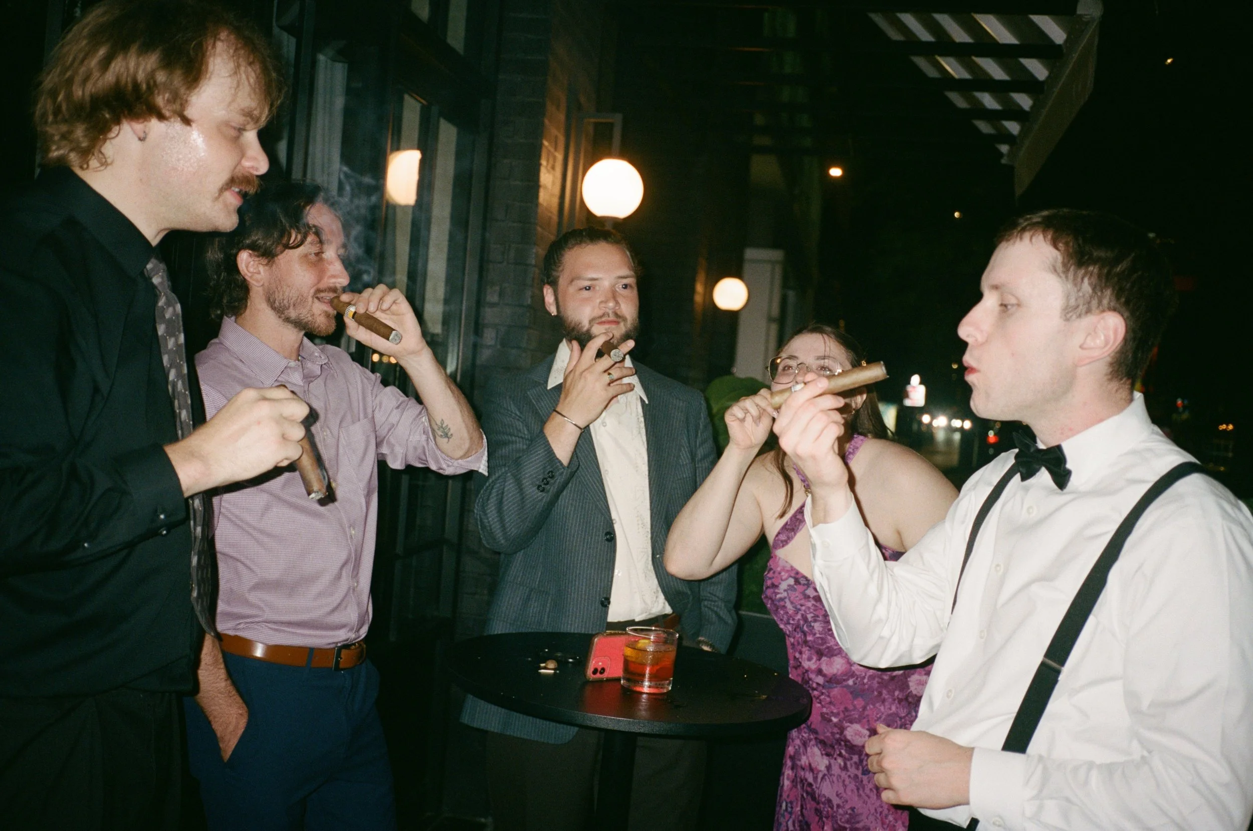 Groomsmen share cigars outside under string lights late in the evening