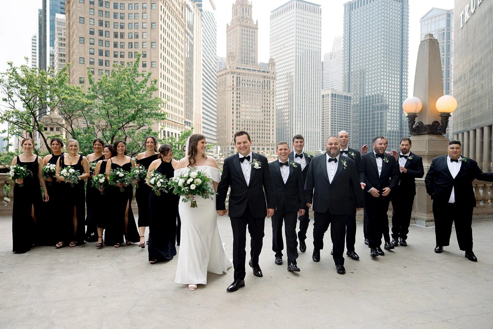 Full wedding party on the Chicago Riverwalk terrace — bridesmaids in black, groomsmen in tuxedos, city skyline behind.