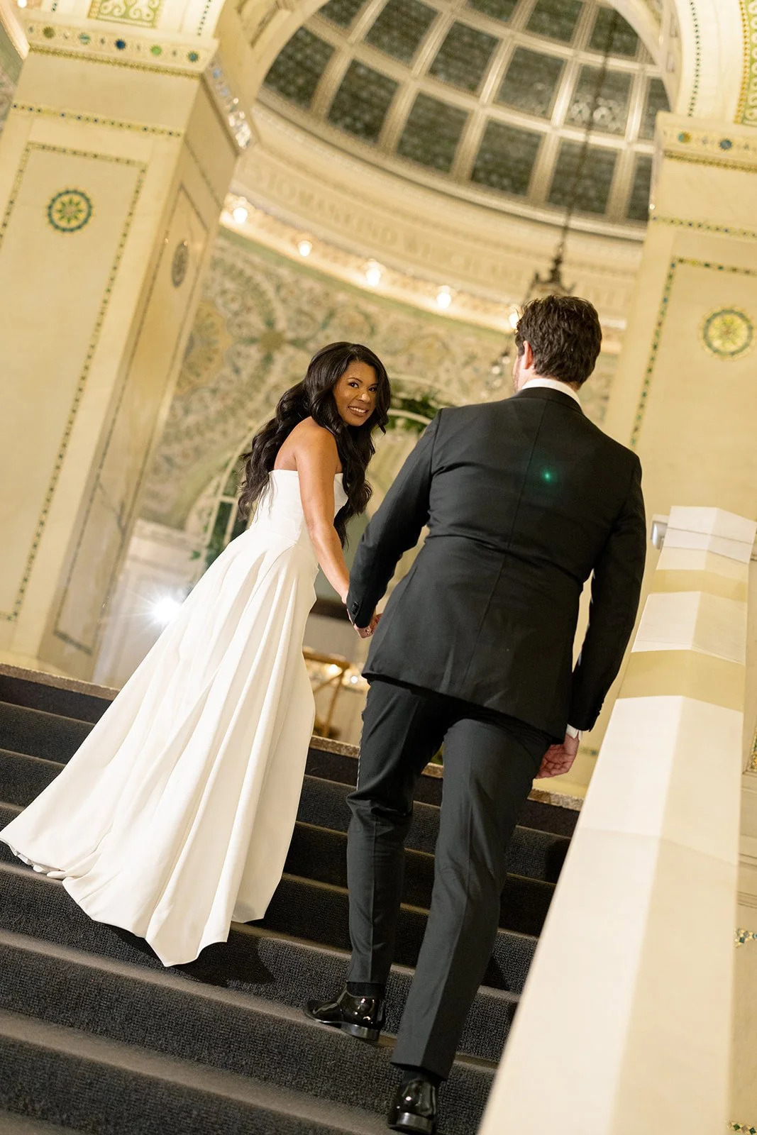 a bride walking her groom up some steps while looking back at the camera