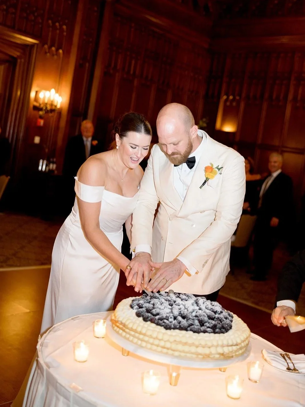 Bride and groom cut their wedding cake together in the University Club Chicago ballroom, candles glowing all around and guests smiling in the background. A warm, candlelit cake-cutting moment — by a Chicago film wedding photographer.
