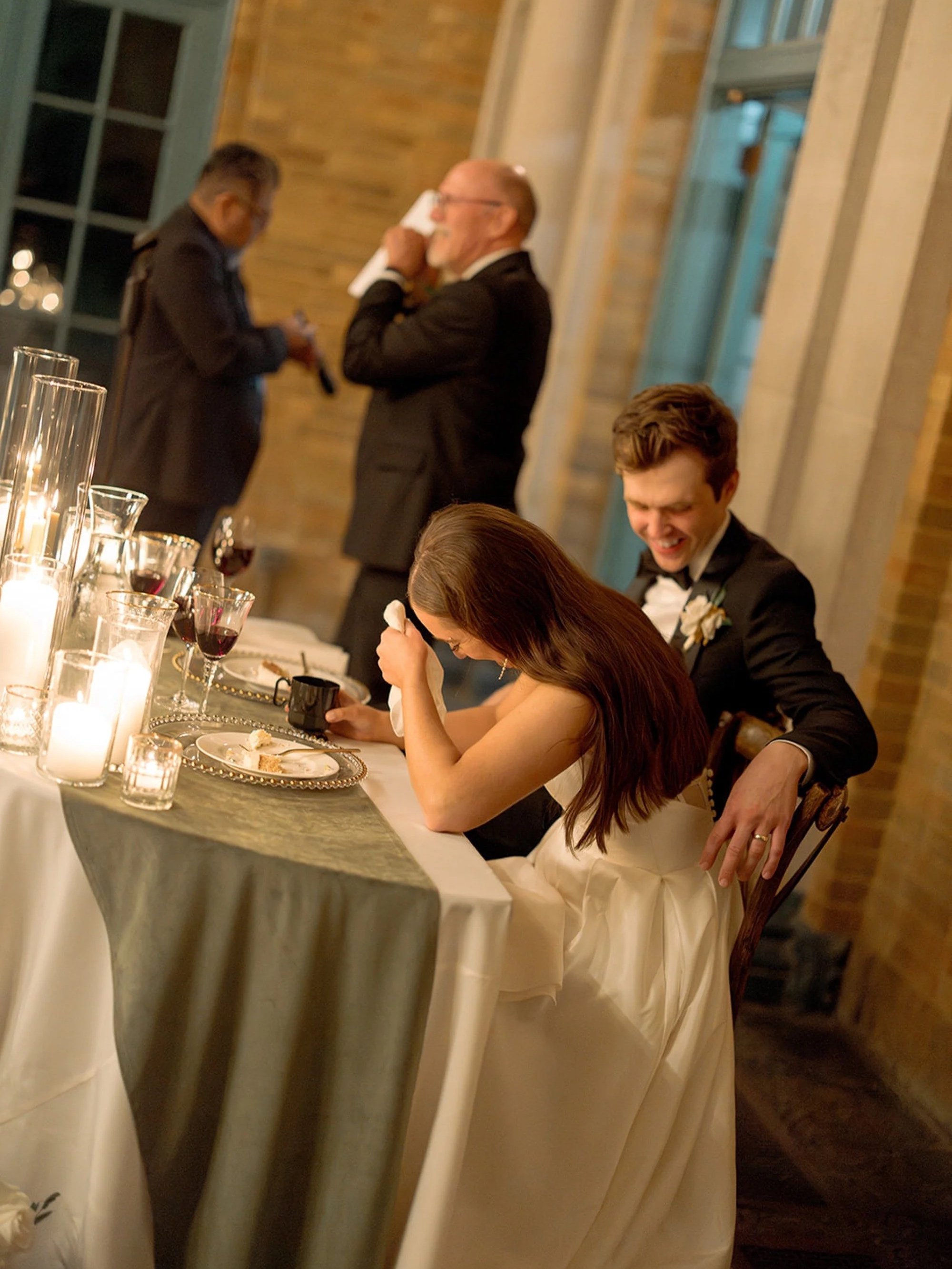 The bride breaks into laughter at the sweetheart table while the groom grins beside her — a perfectly unscripted reception moment captured on film at Columbus Park Refectory