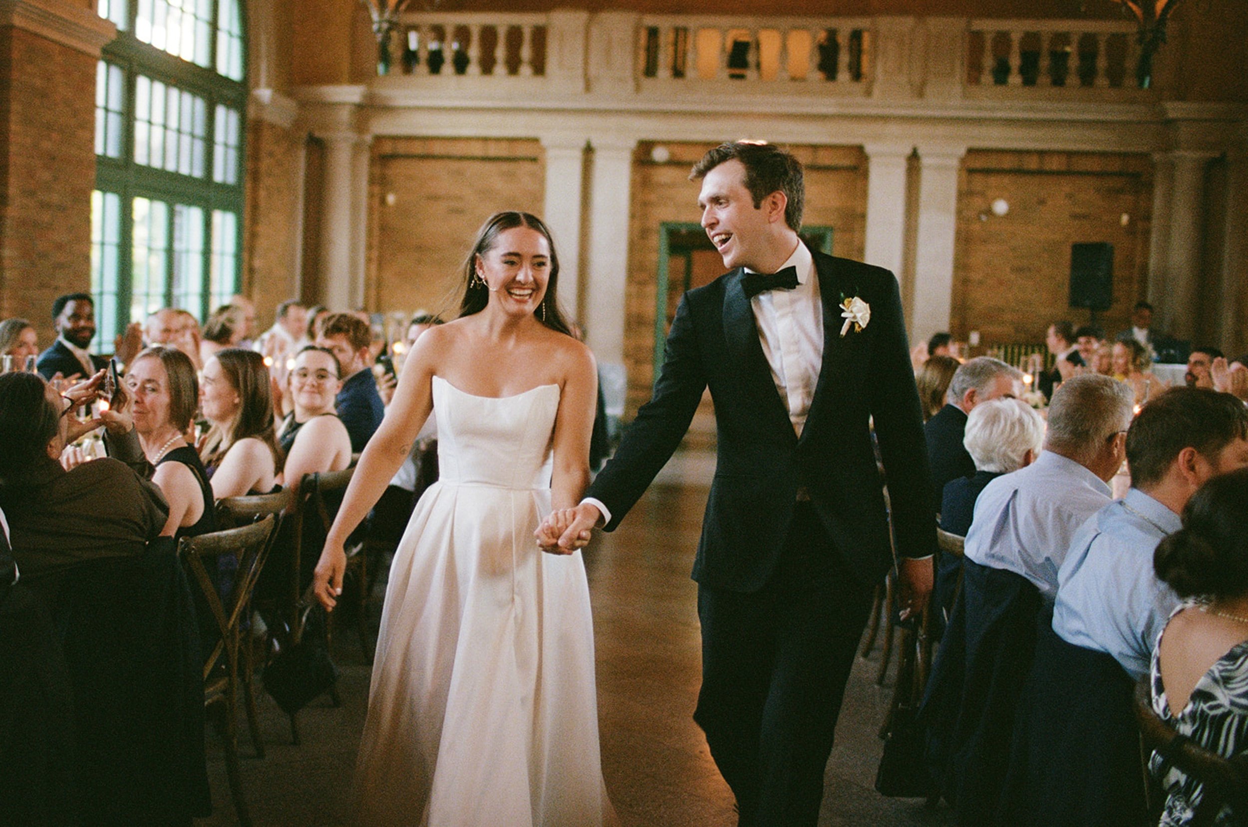 The couple enters their reception to a room full of cheers — hands clasped, smiles unstoppable, the warm glow of the Columbus Park Refectory hall waiting to receive them