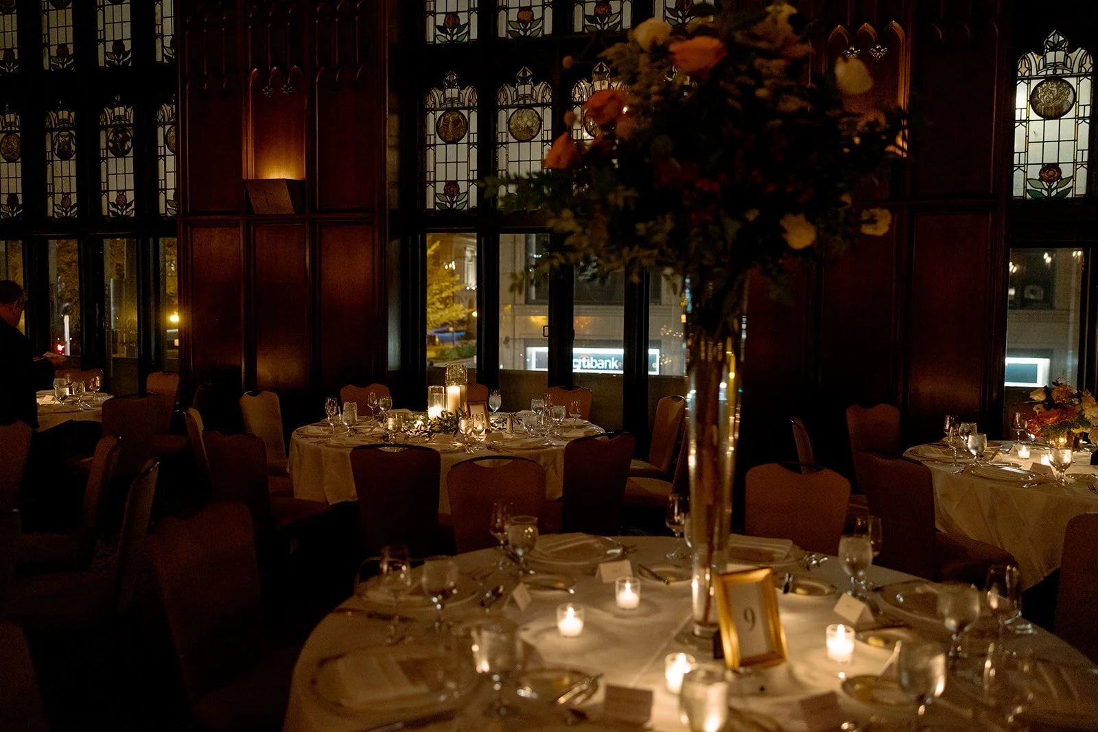 The University Club Chicago reception room fully set for dinner — tall floral centerpieces anchoring each table, candlelight glowing across white linens, stained glass windows presiding in the background.