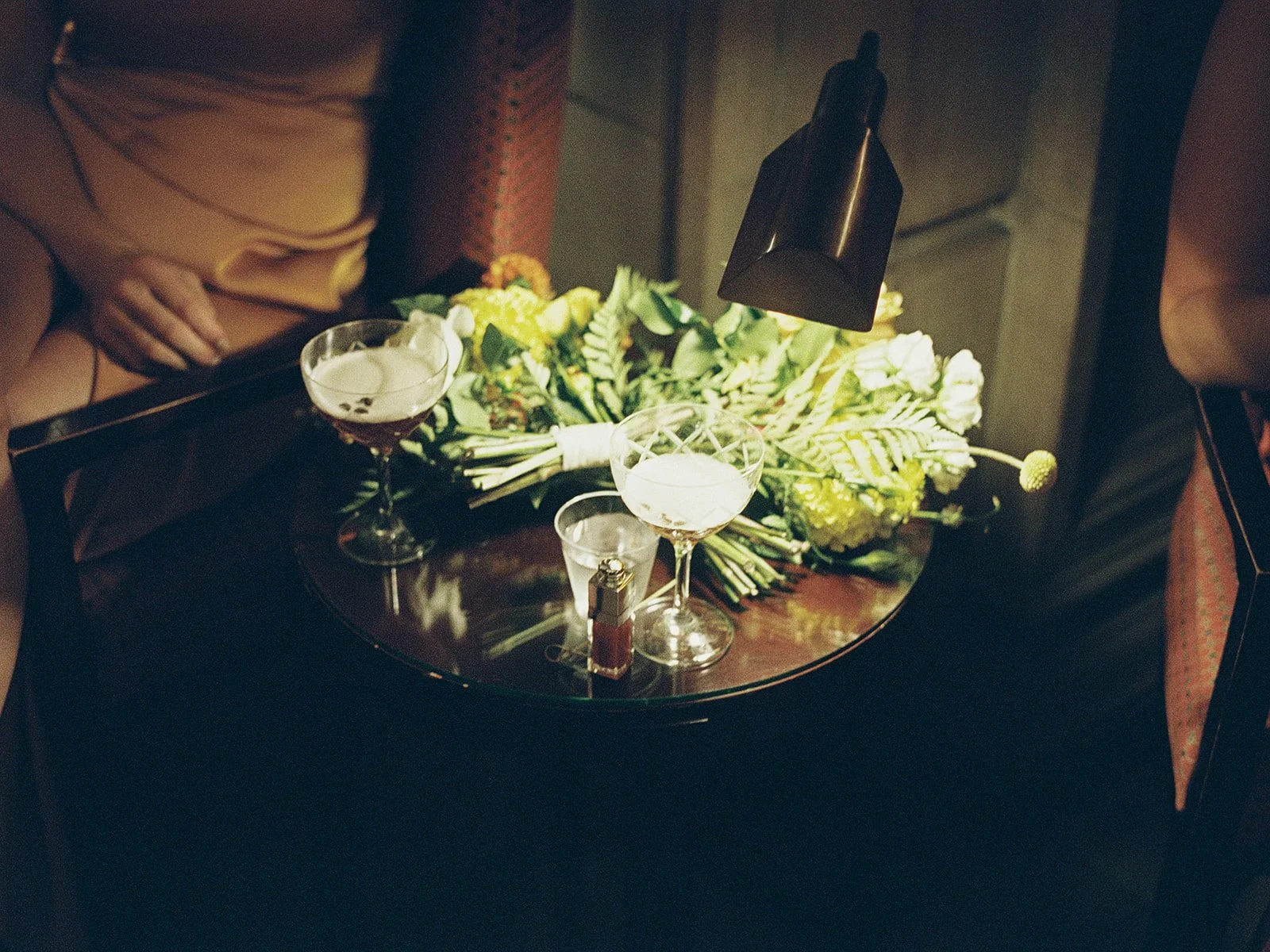 Two cocktail coupes and the bride's lush, green-forward bouquet resting side-by-side on a small table — a still life of the in-between moments. Warm light and film grain make this detail feel like a painting