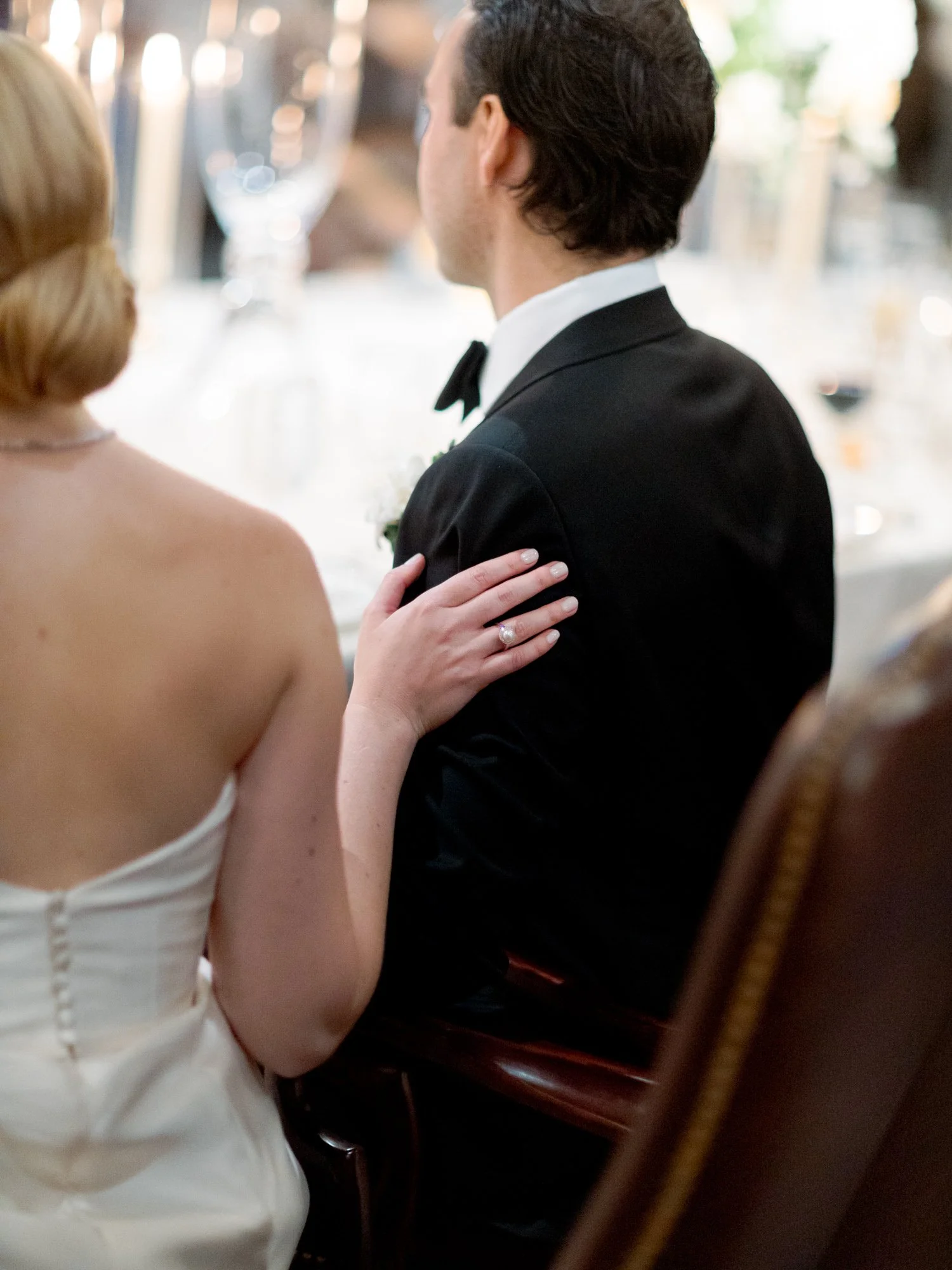 The bride’s hand rests gently on the groom’s back at the sweetheart table, her engagement ring in soft focus, taper candles glowing in the background.
