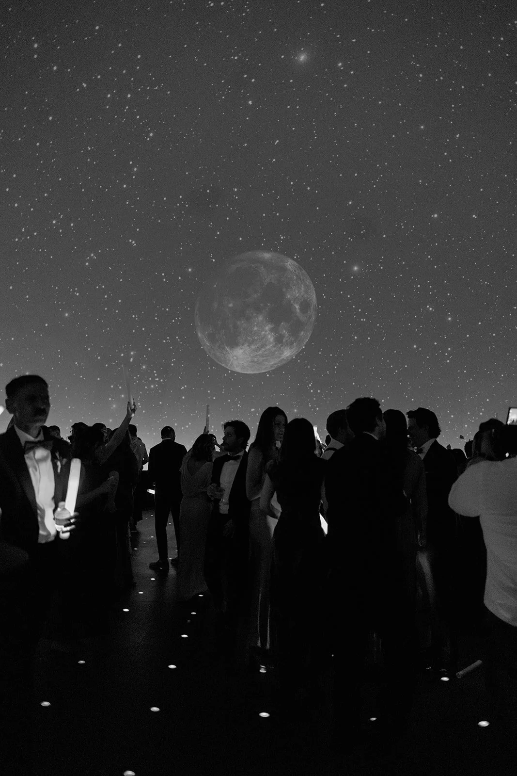 Black and white candid of wedding guests dancing under a projected moon and star-filled sky at an afterparty inside the Adler Planetarium in Chicago, photographed by Louie Abellera.