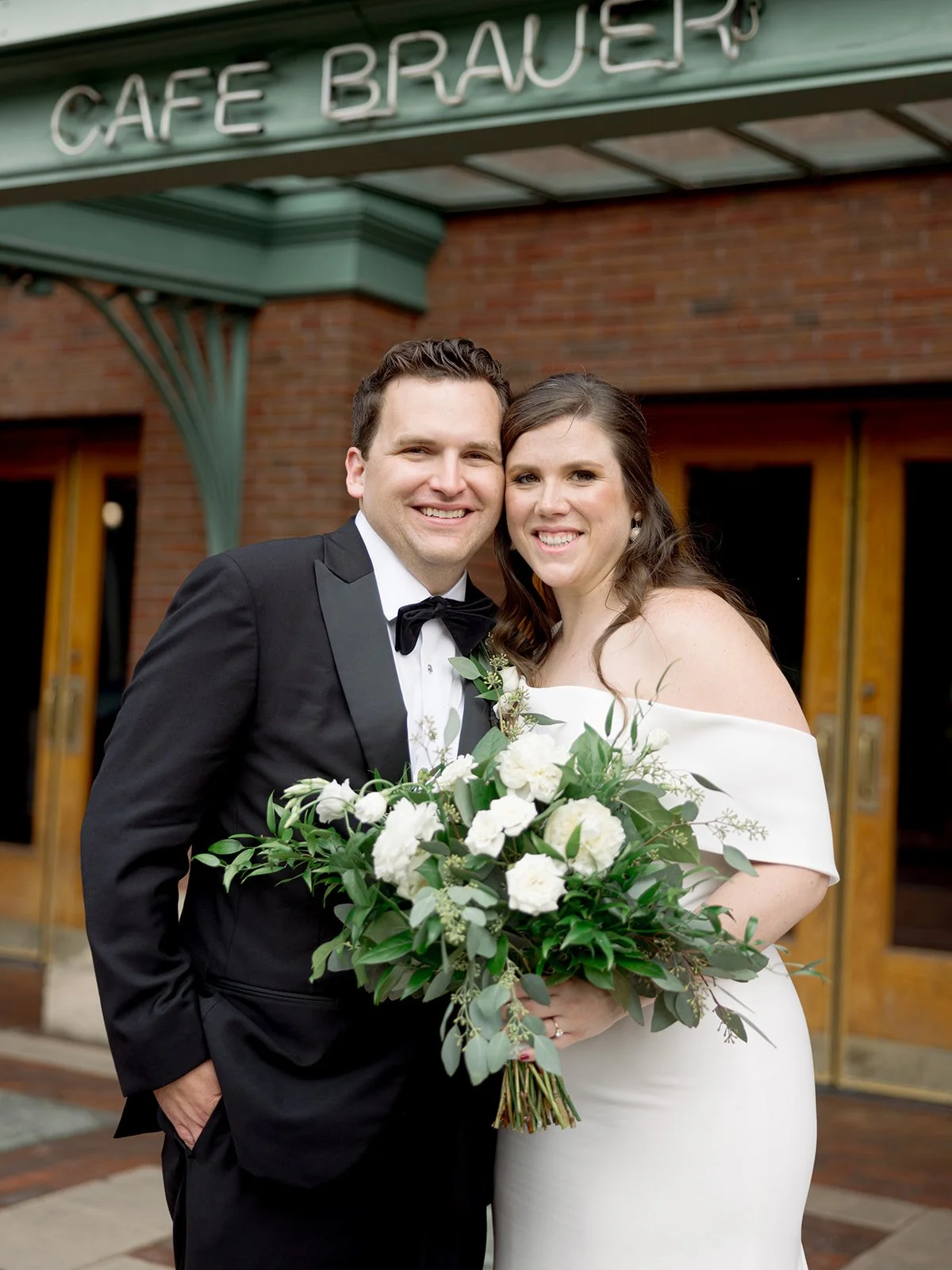 Close, warm portrait of the beaming couple in front of the Cafe Brauer entrance sign in afternoon light.