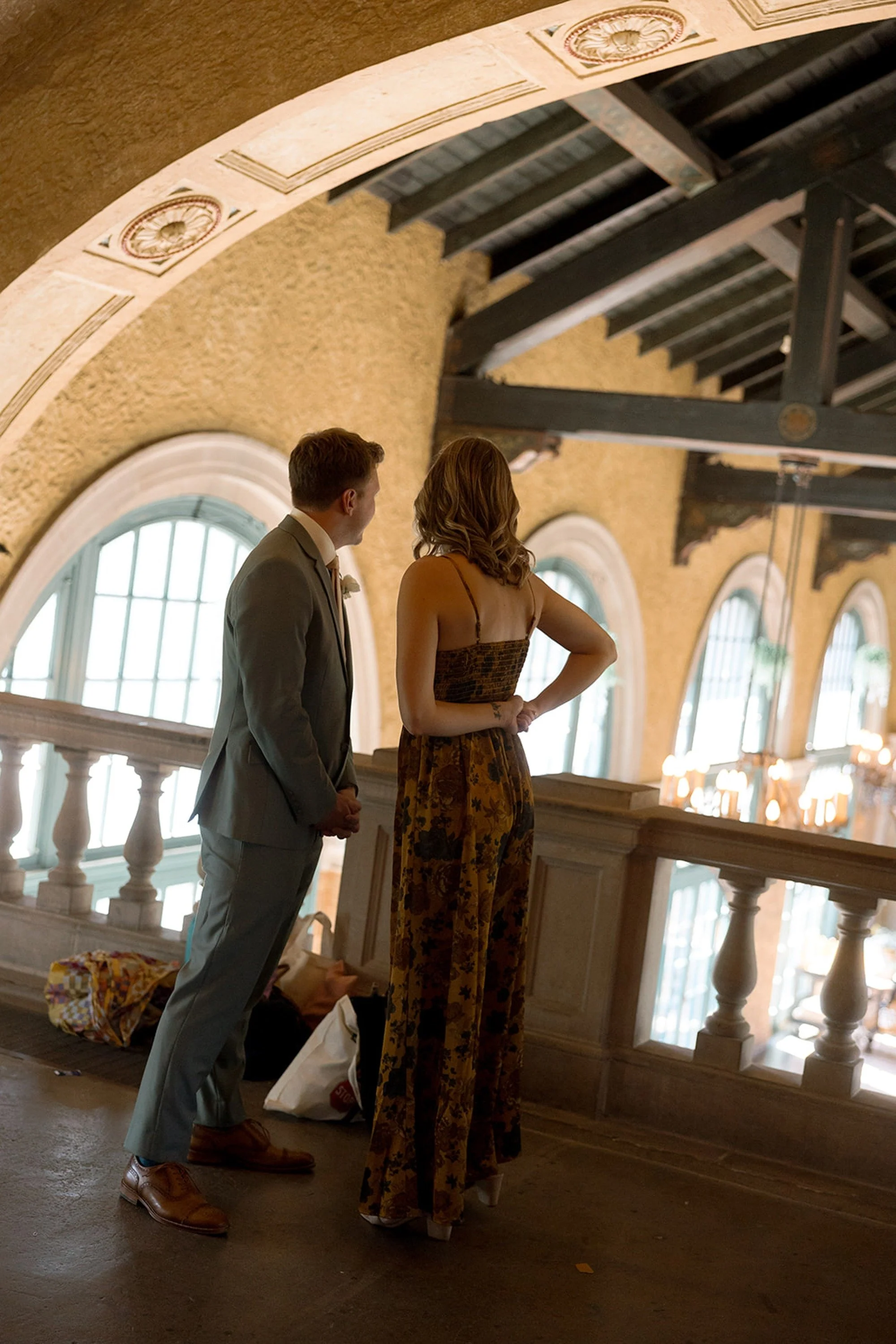 Two guests lean in for a private conversation on the mezzanine balcony of Columbus Park Refectory — the chandelier-lit hall glowing below, arched windows softly lit around them