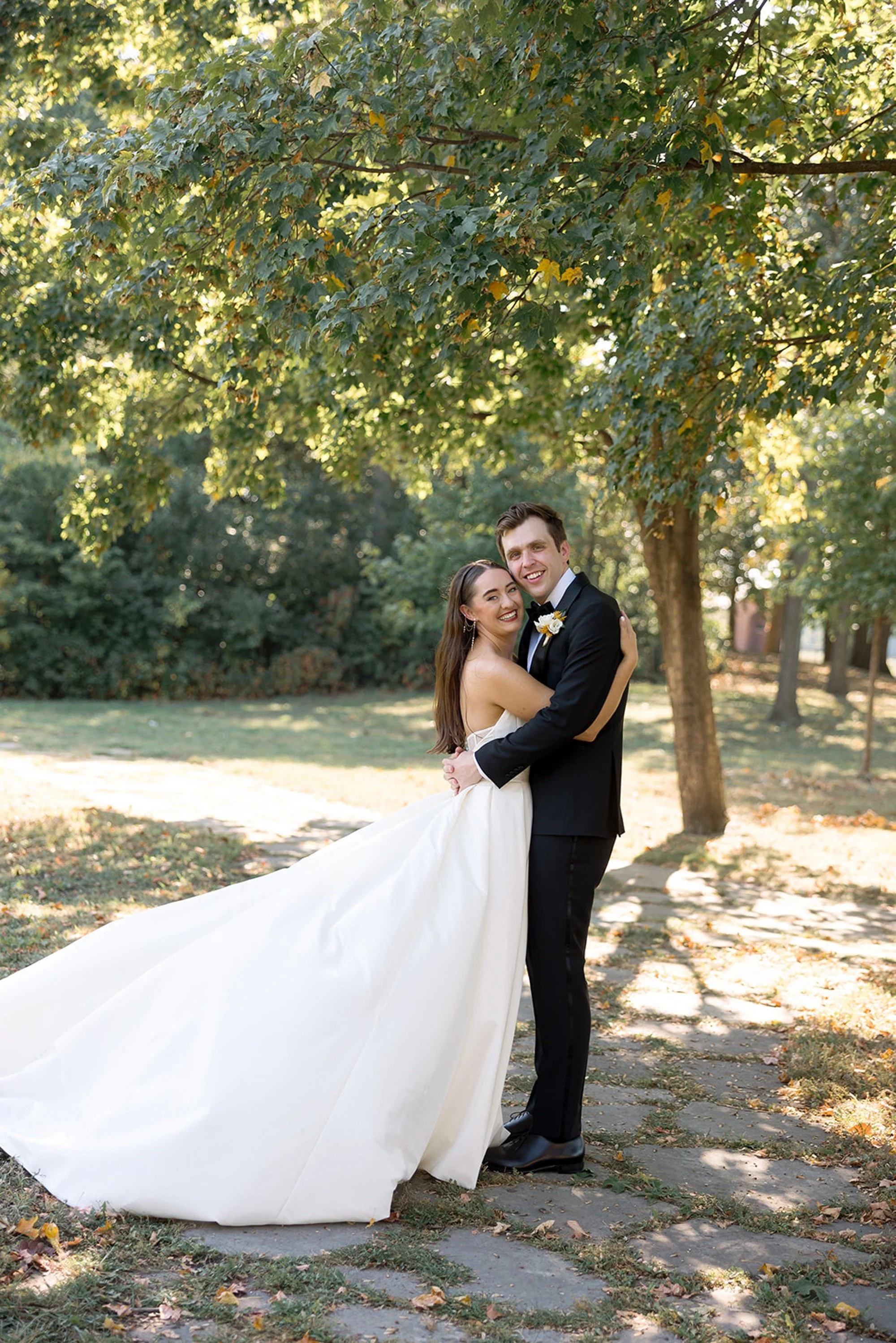 Cradled under a canopy of trees, the couple pulls close — the bride's full ballgown sweeping wide across the stone path, a classic film portrait at Columbus Park Refectory. Chicago film wedding photographer.