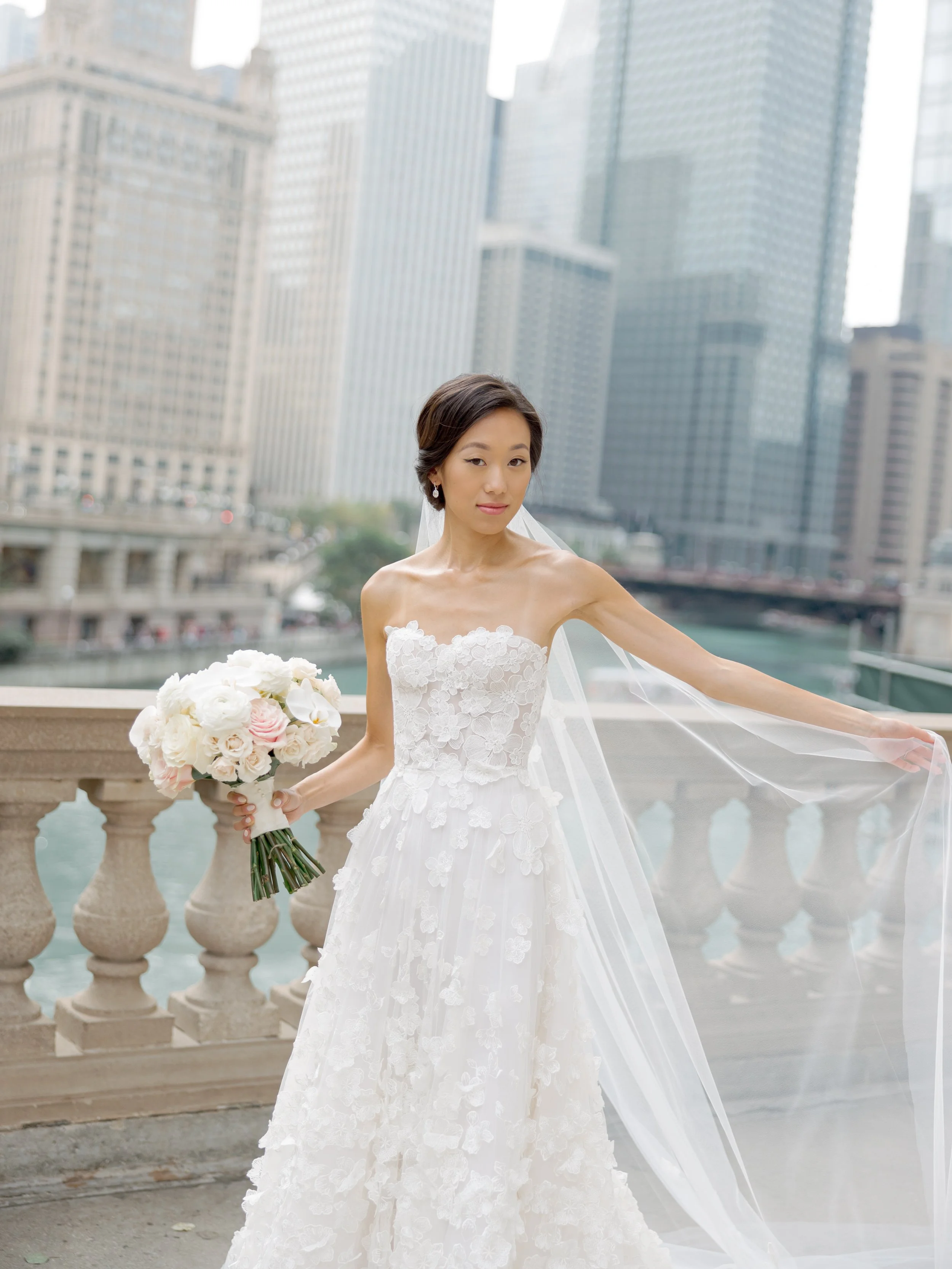 Bride holds her veil and bouquet along the Chicago River with skyscrapers softening in the background, film portrait
