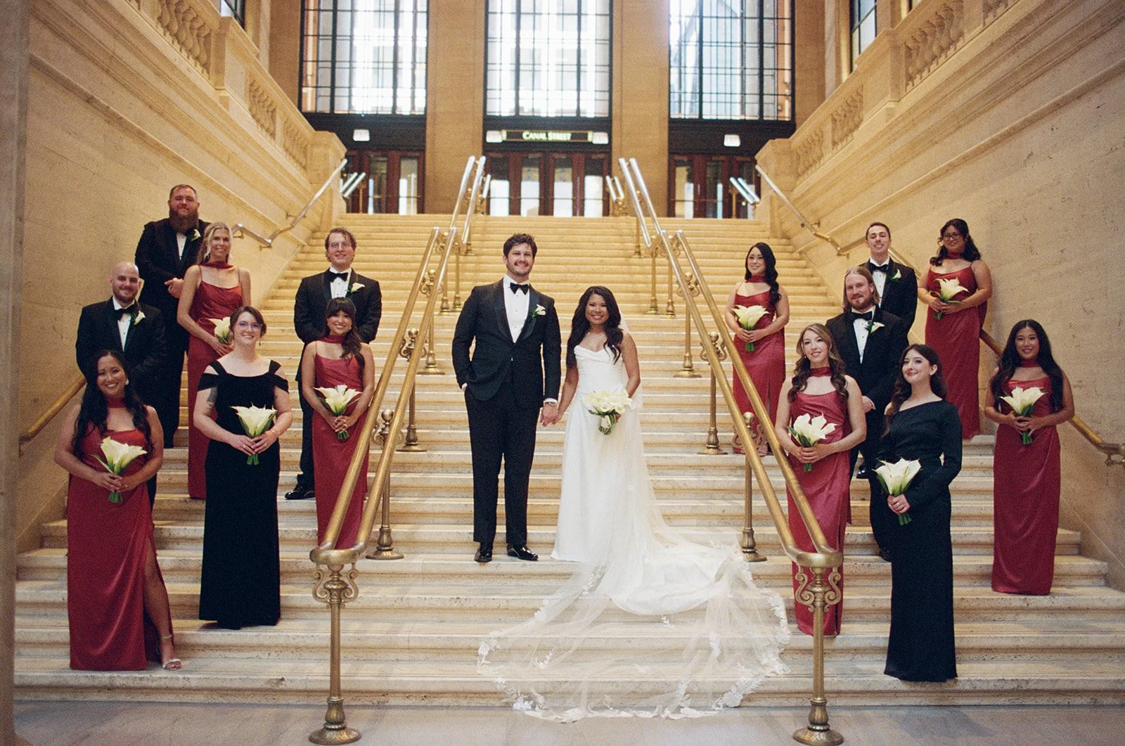 The full wedding party in formal wear — burgundy bridesmaids' gowns and black groomsmen's suits posed on Union Station's grand staircase. Shot on film by a Chicago wedding photographer.