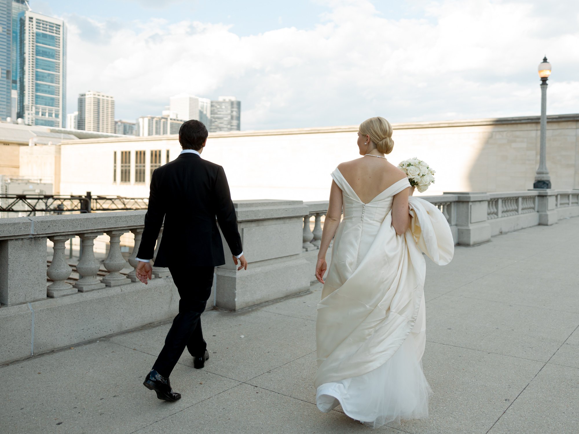 Bride and groom walk hand-in-hand away from the camera across a neoclassical stone terrace, the full Chicago skyline rising at the horizon behind them.