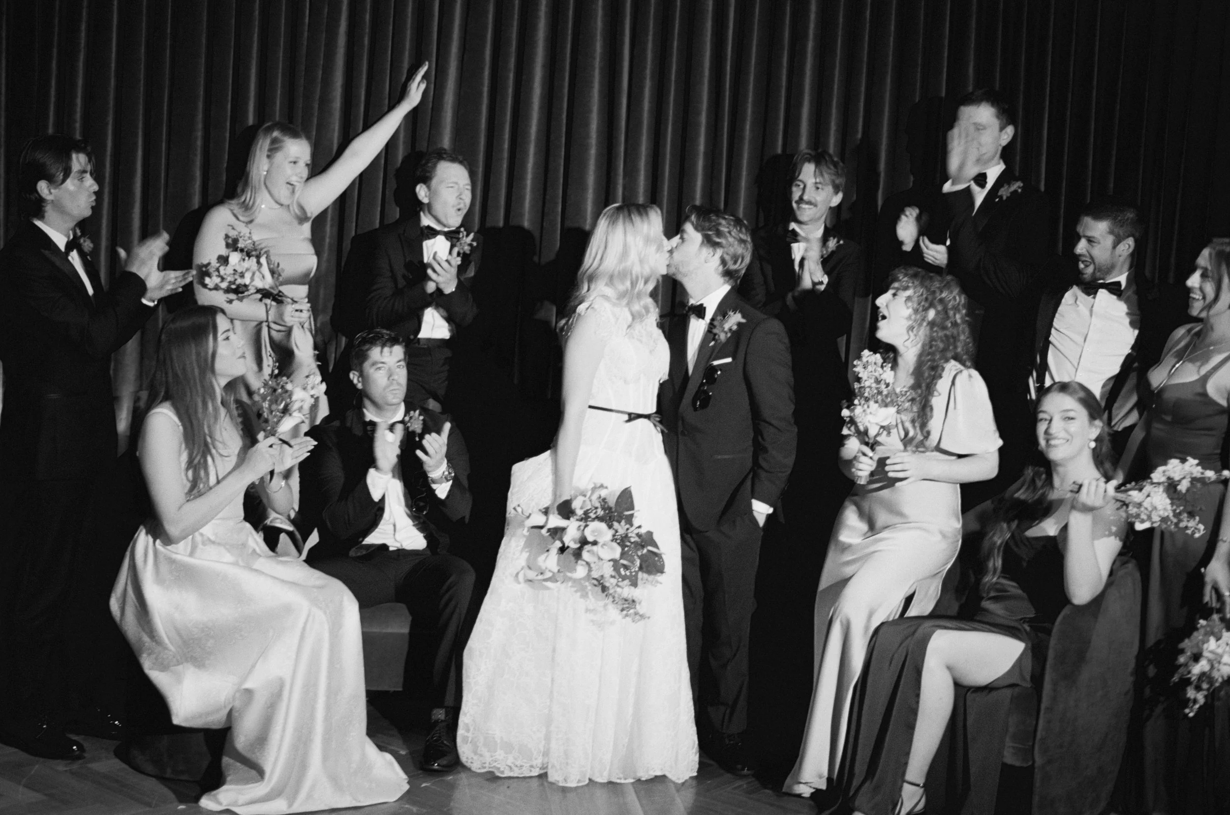 Black and white photo of guests cheering as the newlyweds kiss inside The Wellsley's red velvet lounge