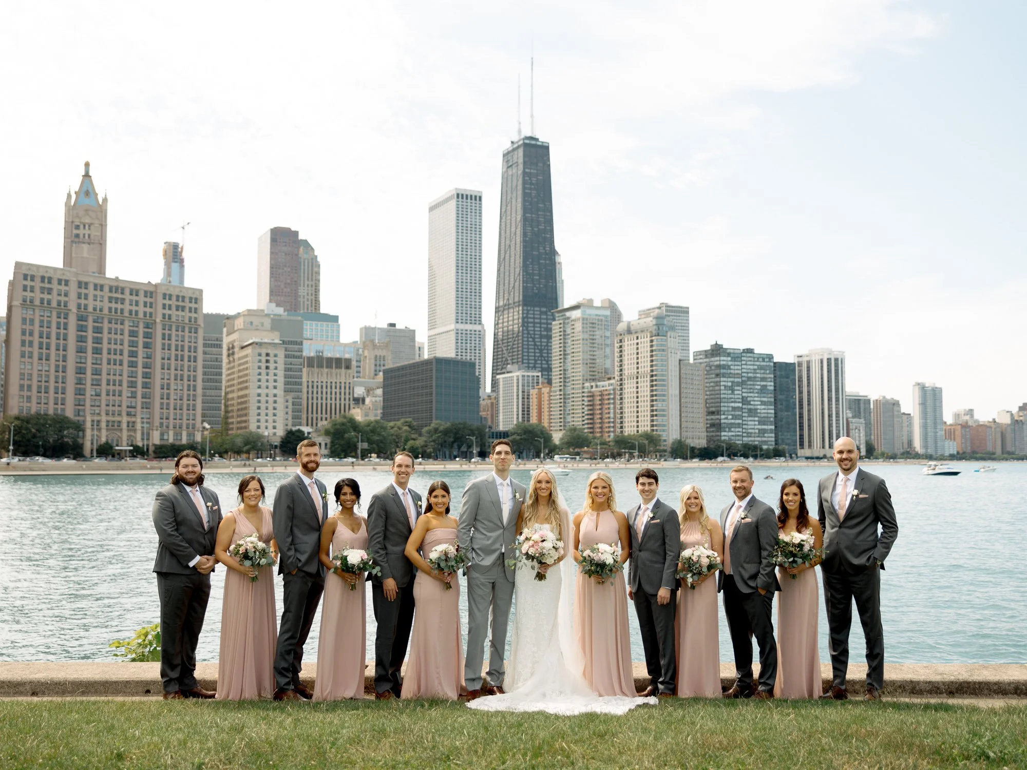 The full wedding party poses at Olive Park with the stunning Chicago skyline and Lake Michigan behind them, bridesmaids in blush and groomsmen in grey.
