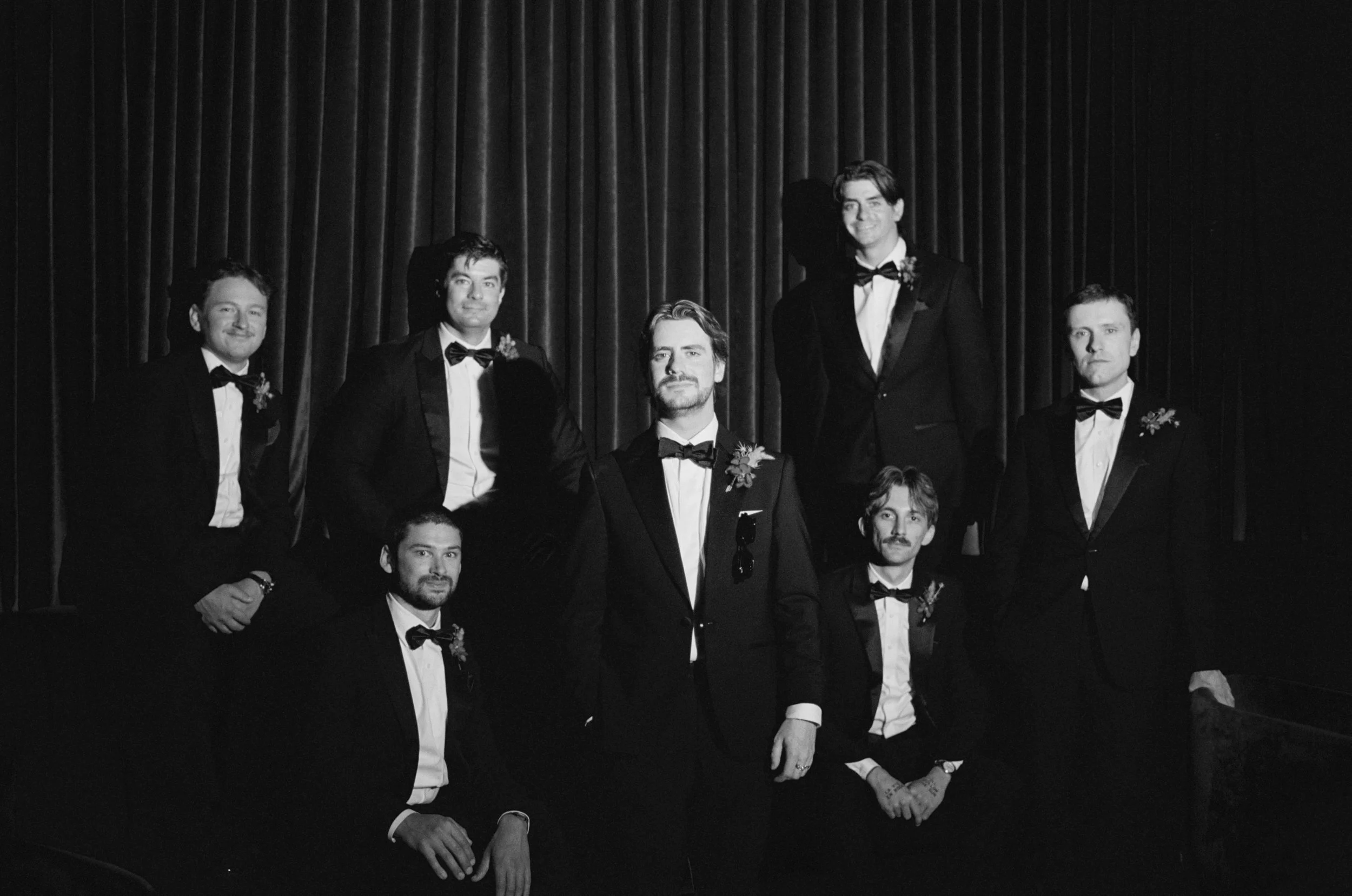 Black and white formal portrait of the groomsmen in tuxedos inside The Wellsley's red velvet lounge