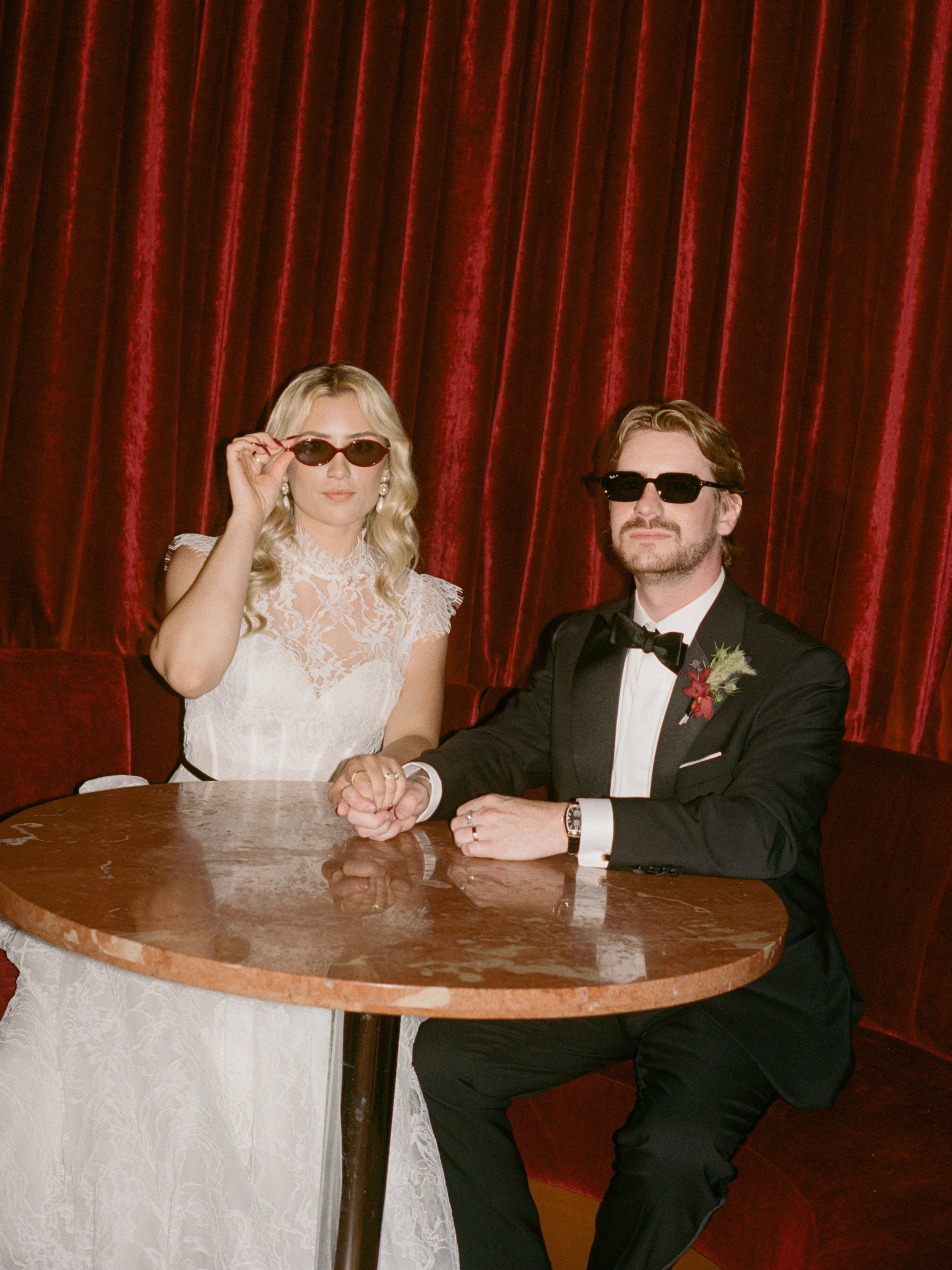 Bride and groom hold hands in sunglasses at their round table portrait in The Wellsley's red velvet lounge