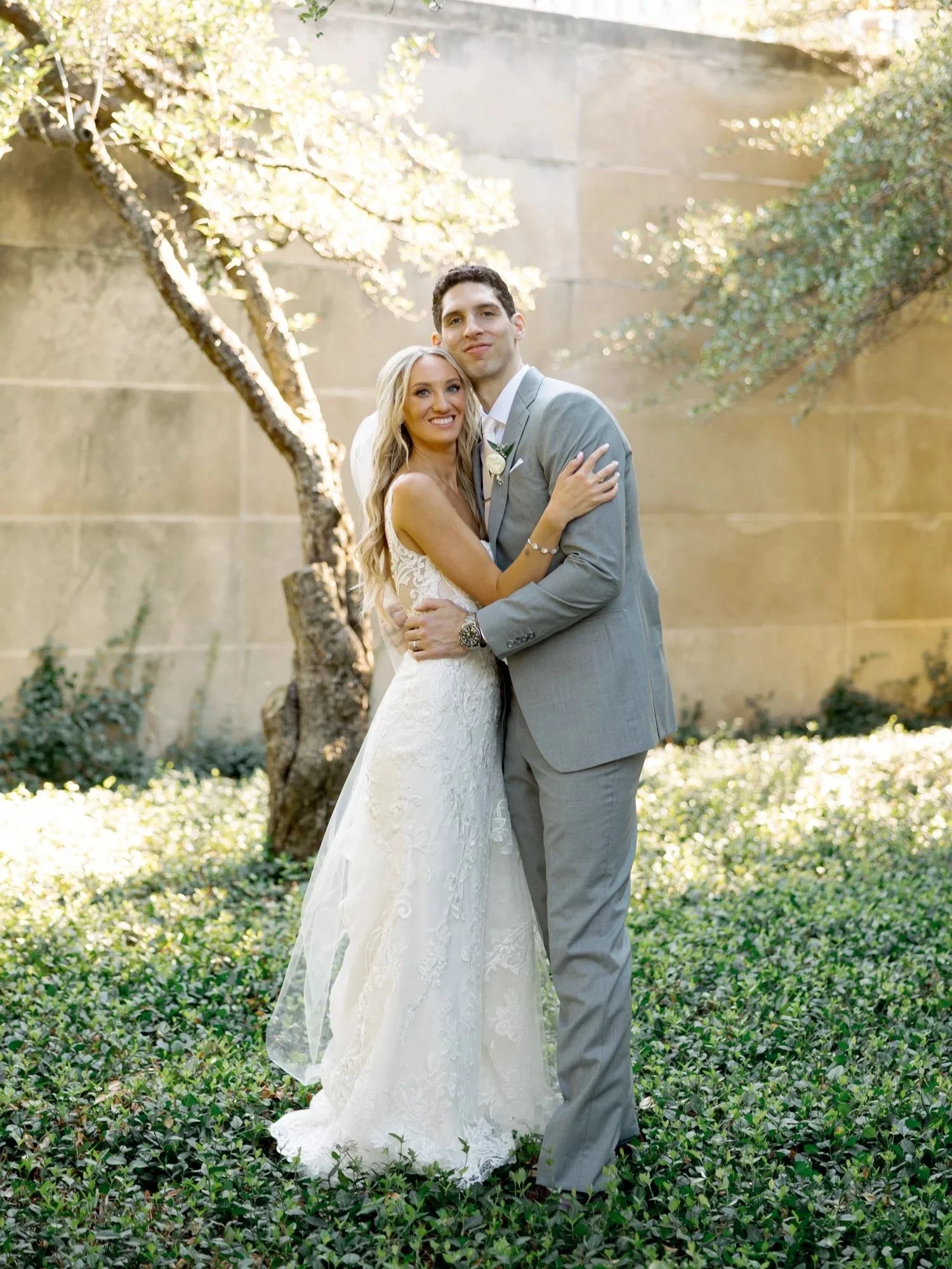 The bride and groom smile warmly in each other's arms in the Art Institute South Gardens, dappled sunlight filtering through olive trees and a stone wall behind.