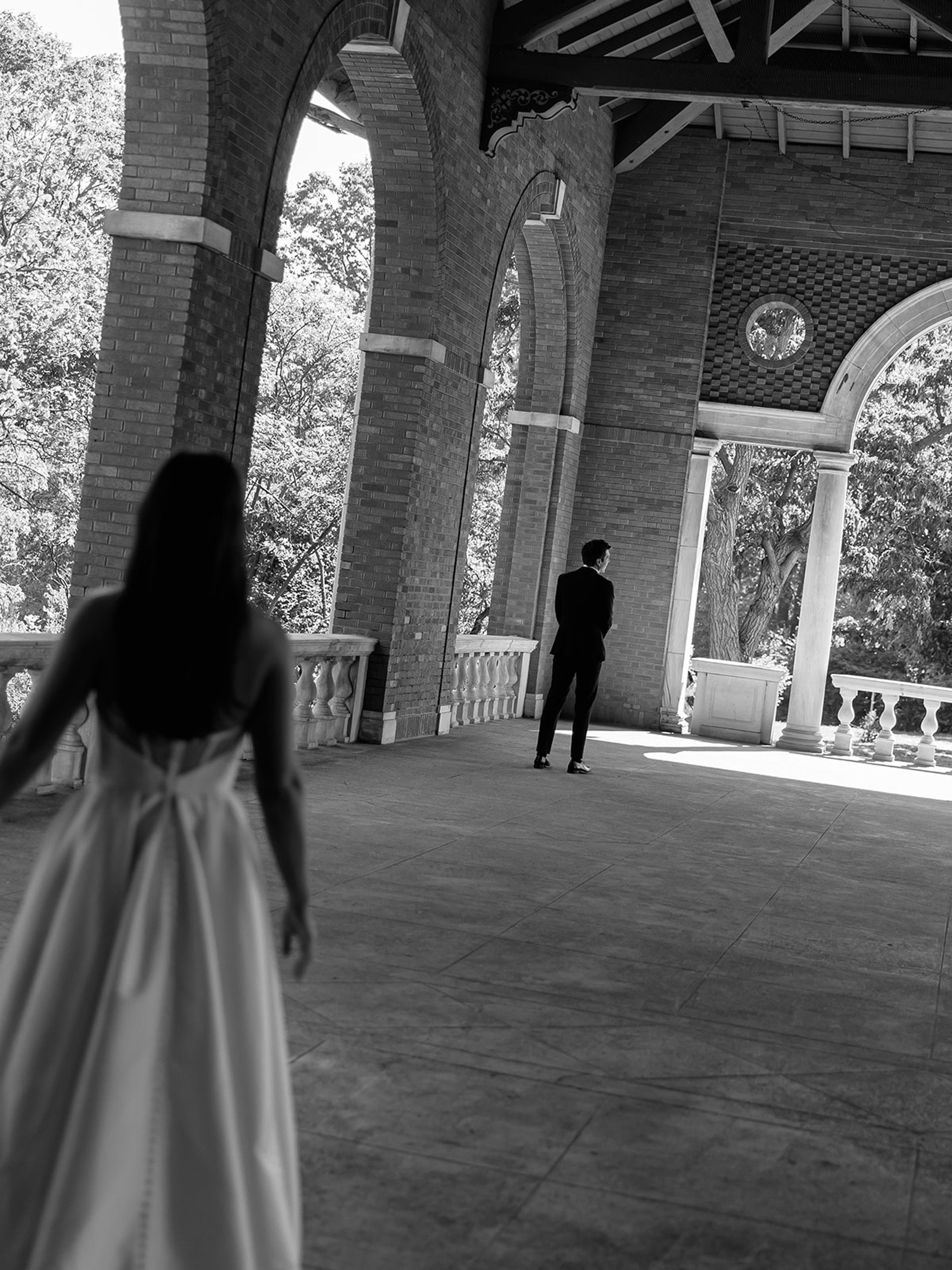 Dramatic and cinematic — a black-and-white wide shot of the bride walking toward her groom, who stands alone at the far end of the Columbus Park Refectory pavilion. The space between them, about to close forever