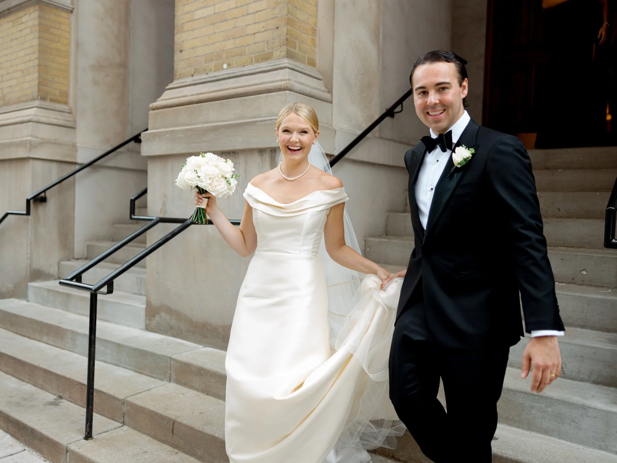 Bride lifts her bouquet and laughs as she and the groom descend stone church steps together, newly married and beaming — shot by a Chicago film photographer.