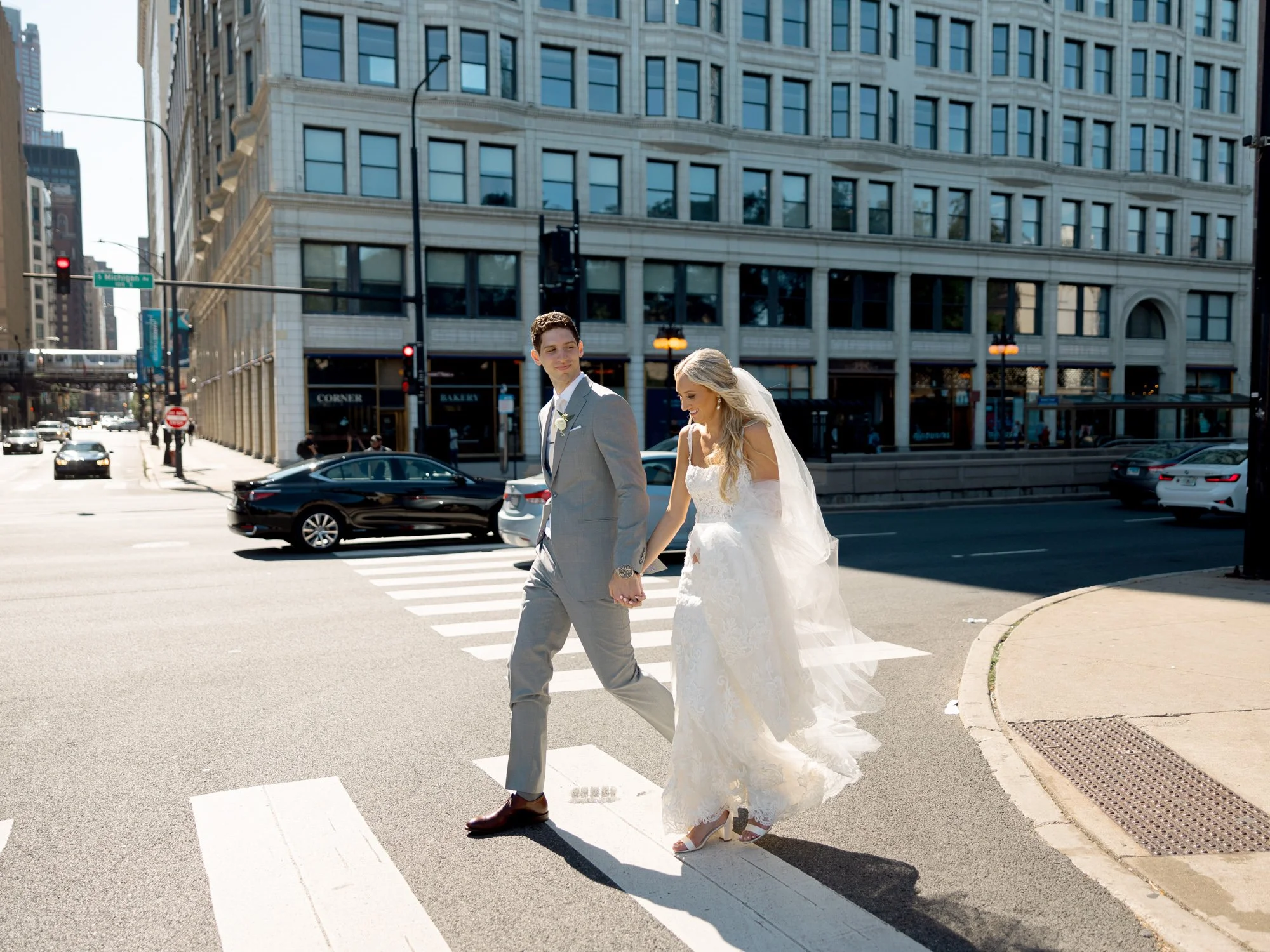 The bride and groom stride hand-in-hand across a sunny downtown Chicago crosswalk, her lace gown and veil flowing gracefully behind them in the breeze.