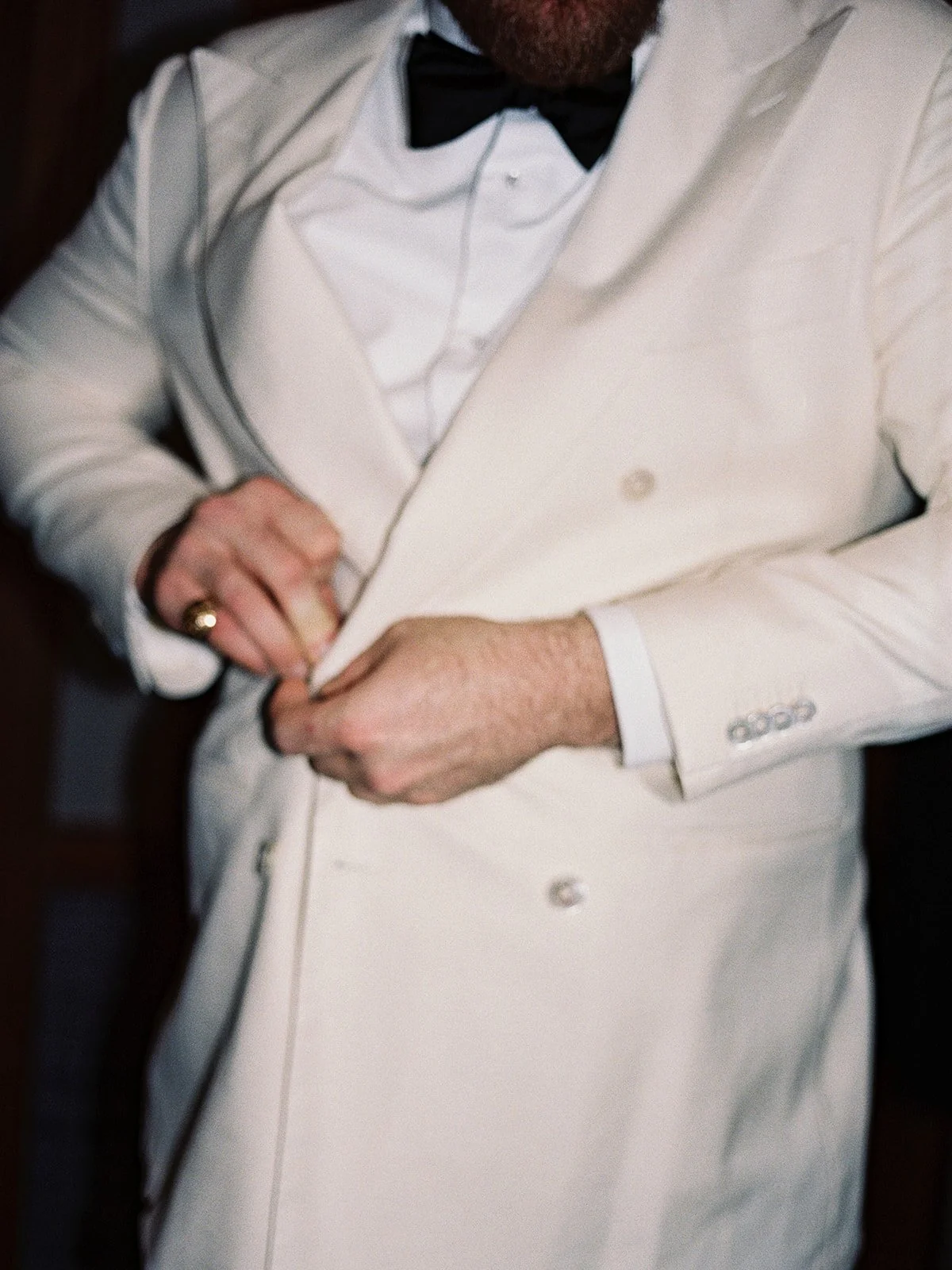 A close-up getting-ready detail: the groom's hands buttoning his white double-breasted dinner jacket, gold signet ring on his finger, bow tie already in place. Film grain gives this quiet moment its texture — by a Chicago film wedding photographer.
