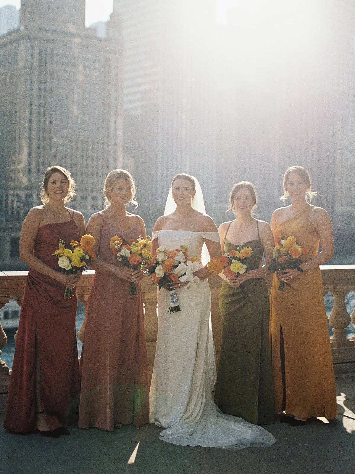 Five women on the Chicago Riverwalk bridge, sun blazing behind them — the bride in white at center, bridesmaids in their earth-tone gowns, each holding a burst of colorful blooms. Backlit, golden, and completely cinematic