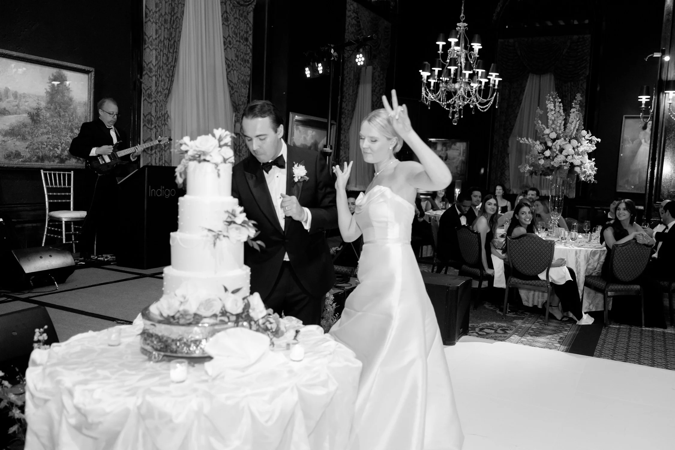The bride throws up a double peace sign beside a towering four-tier floral wedding cake in the Union League Club ballroom — playful, perfect, on-brand.