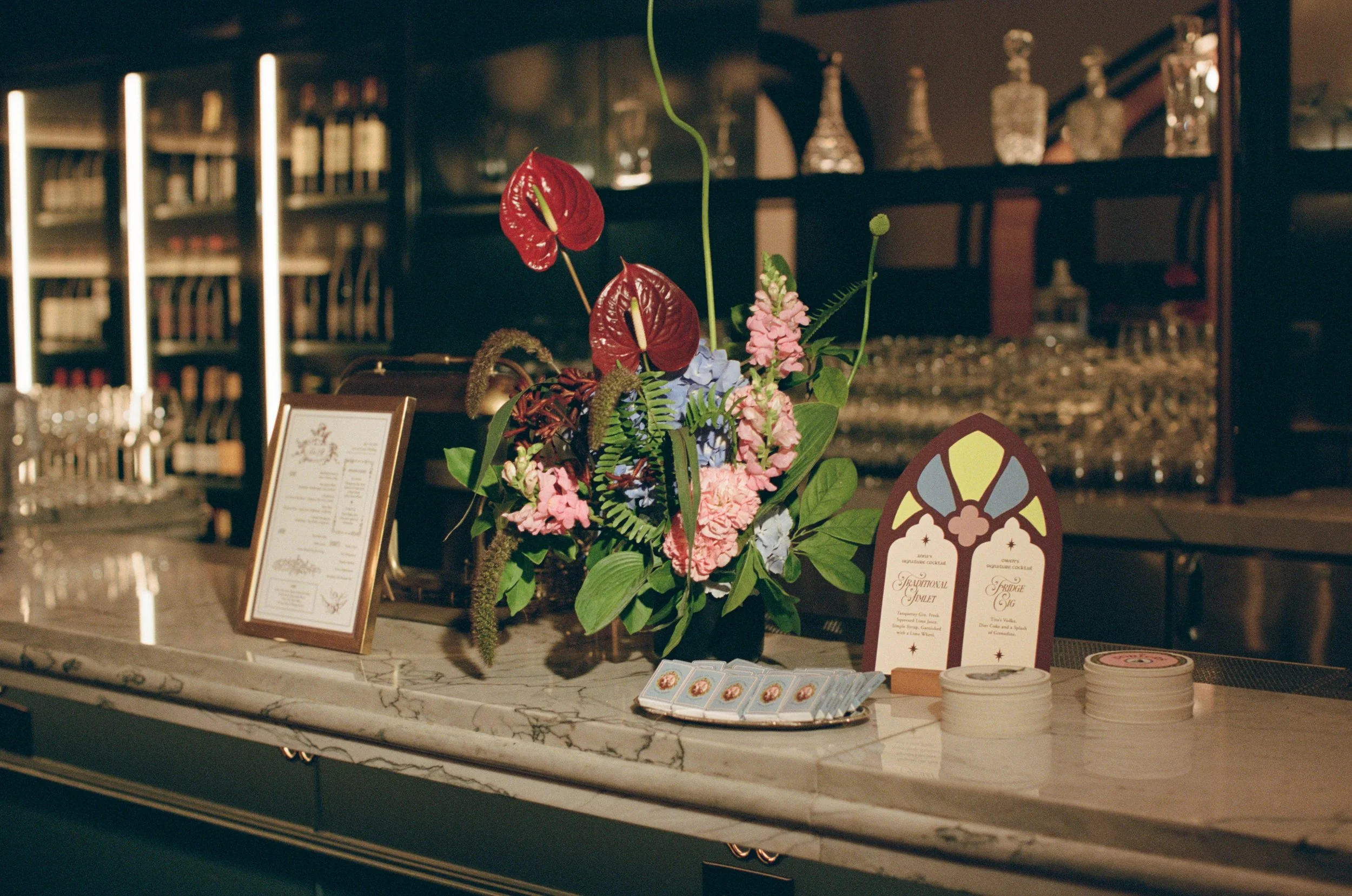Bar counter styled with red anthurium florals, a welcome sign, and glassware at The Wellsley