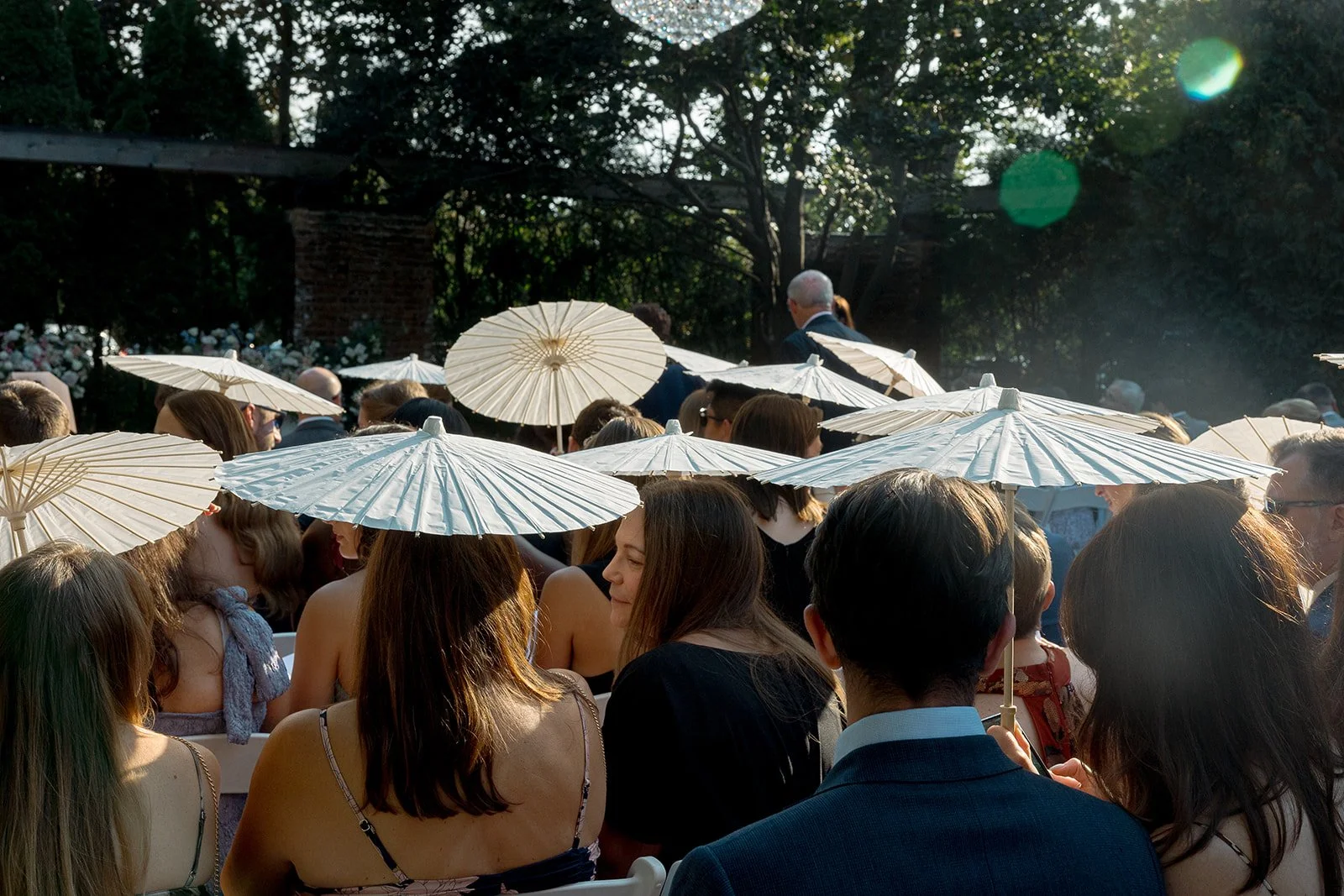 Candid shot of wedding guests holding white parasols during a sun-drenched outdoor ceremony at Community House Winnetka, photographed by Chicago film wedding photographer Louie Abellera.