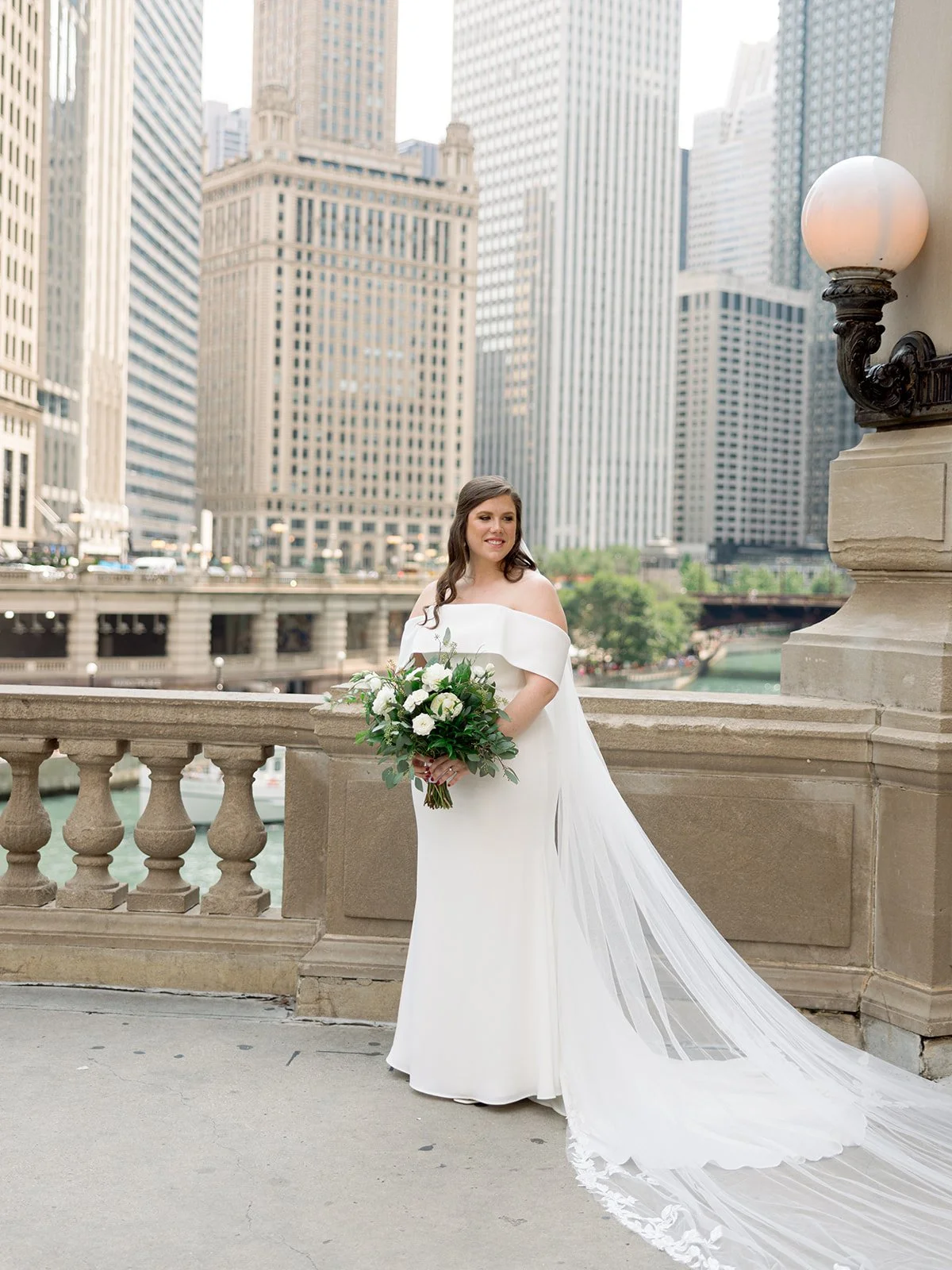Bride standing on a stone balustrade overlooking the Chicago River, cathedral lace veil trailing, city skyline behind her.