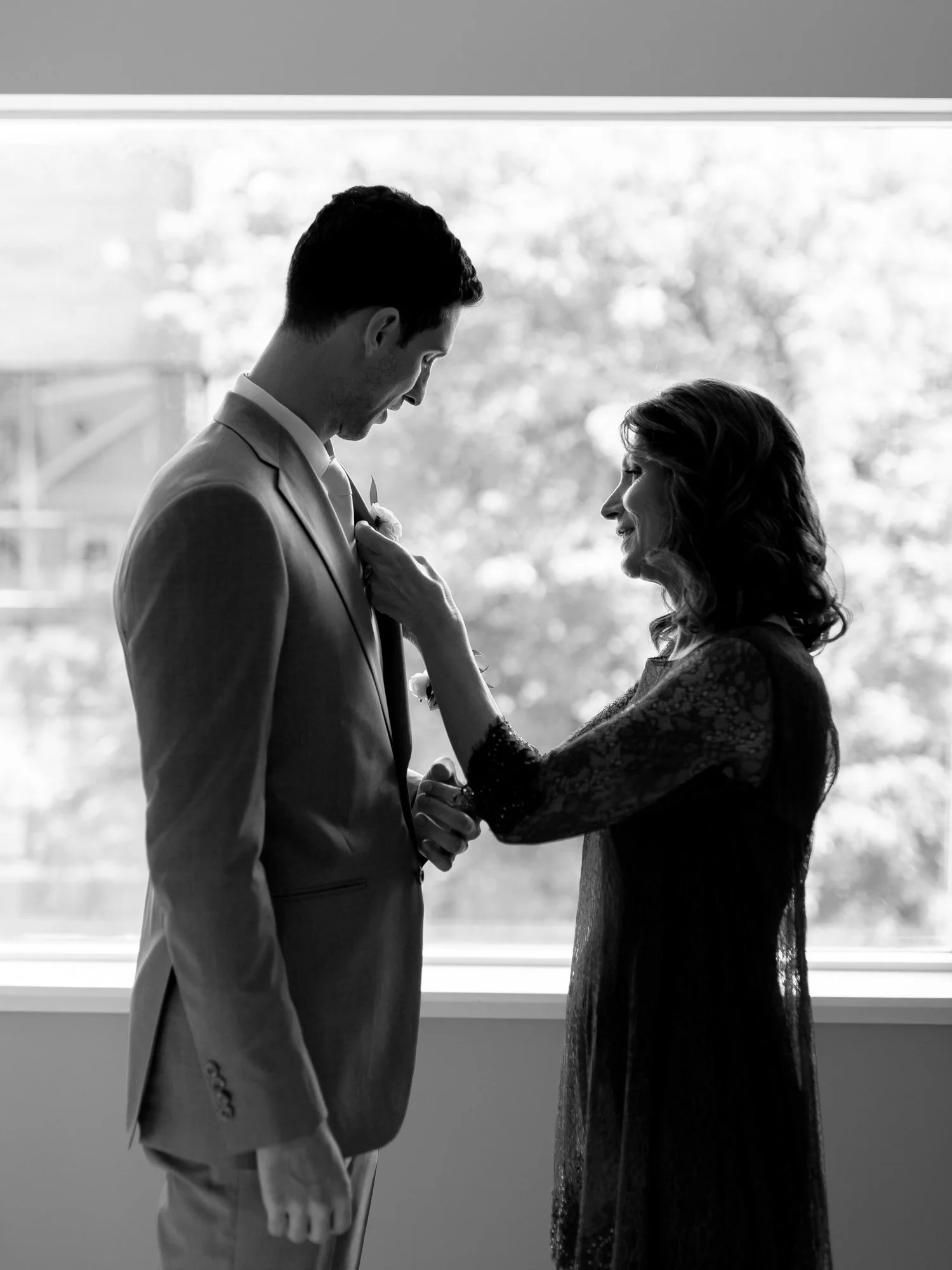 A touching black-and-white silhouette of the groom's mother pinning his boutonniere by a bright window at the Chicago Illuminating Company before the ceremony.