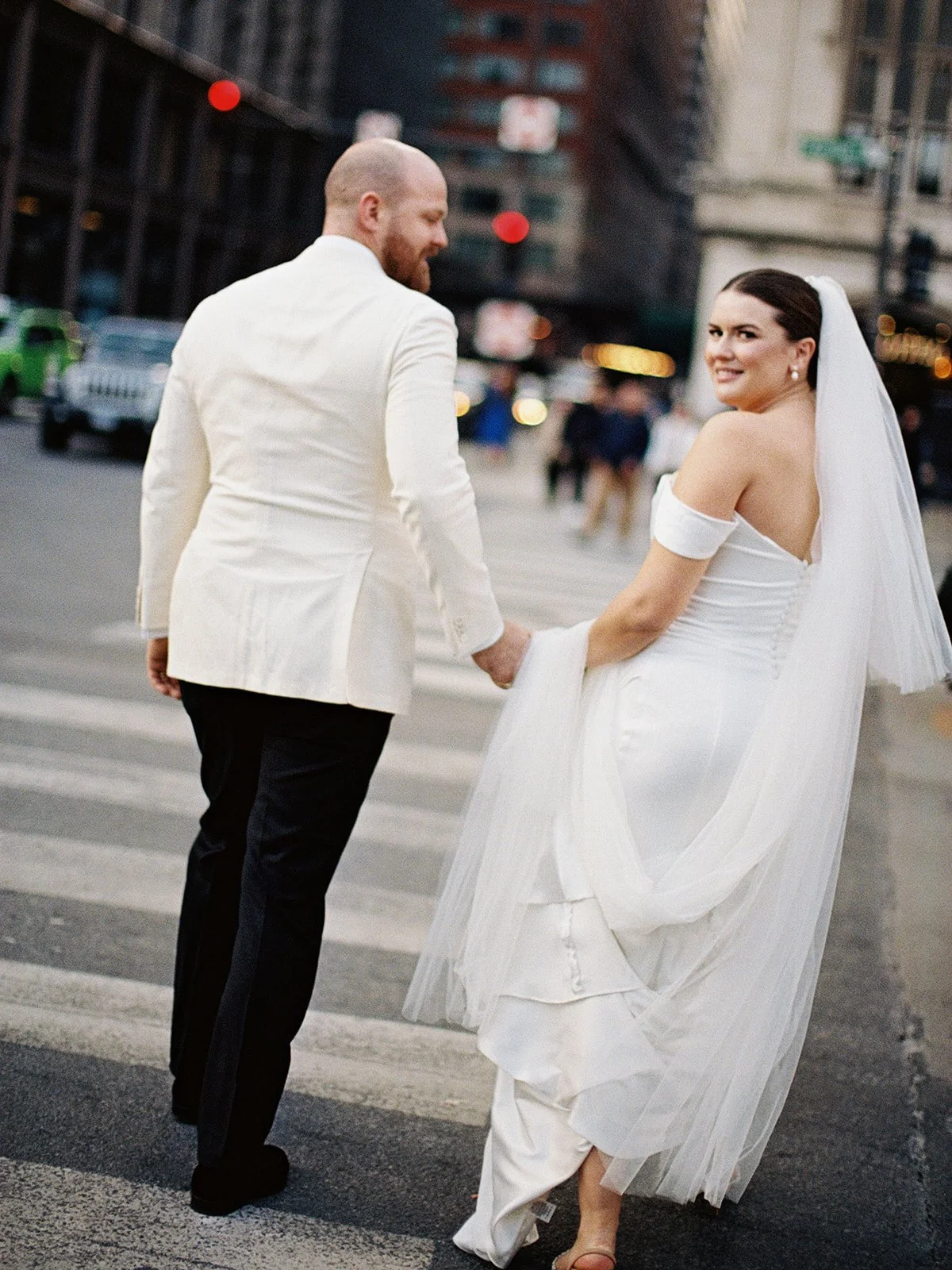 The couple crosses a downtown Chicago intersection hand-in-hand, the bride glancing back over her shoulder with a knowing smile — the city in motion all around them.