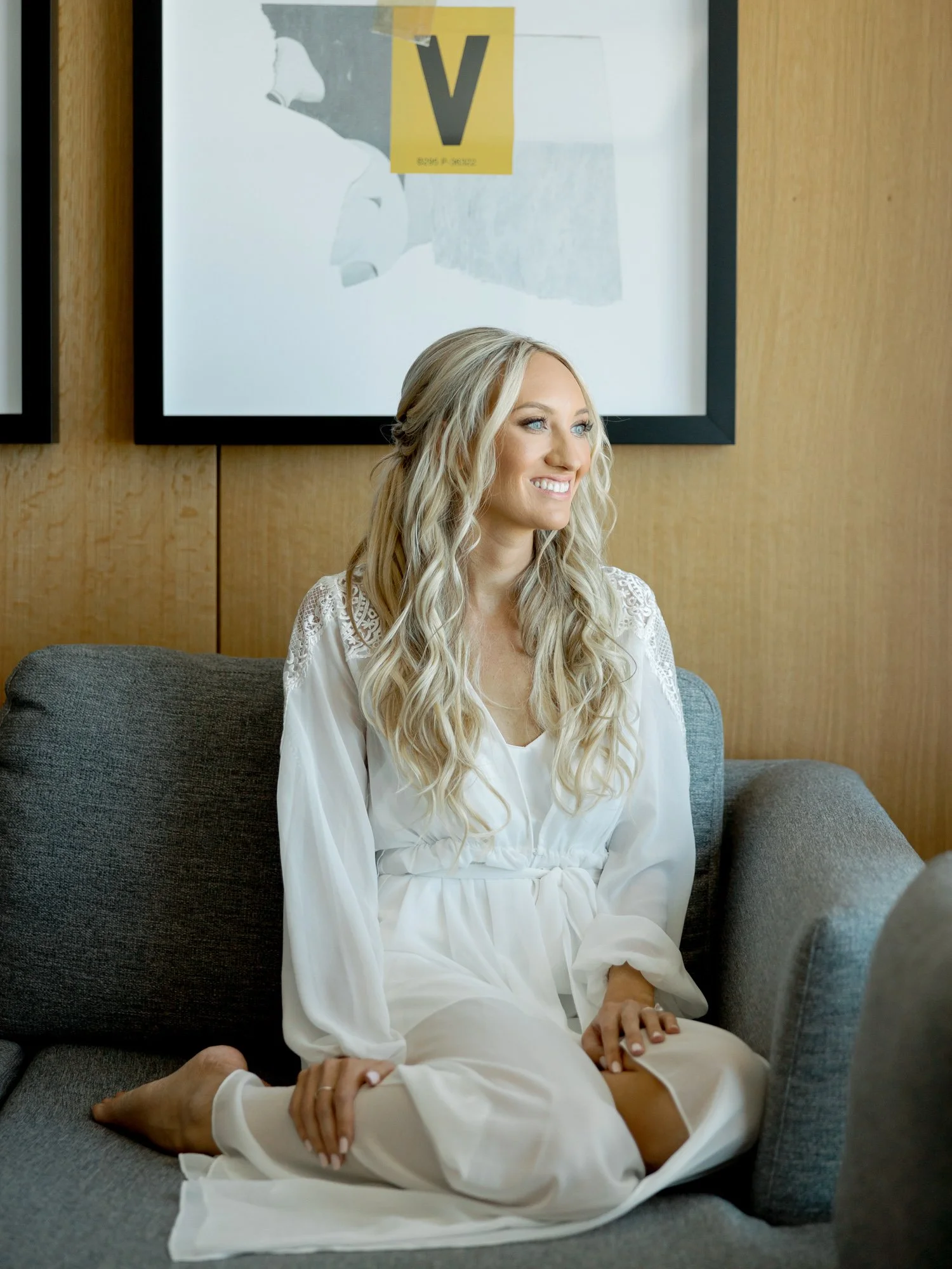 The bride relaxes on a modern hotel sofa in a flowing white robe, her effortless smile and loose wavy hair radiating calm joy on her wedding morning.