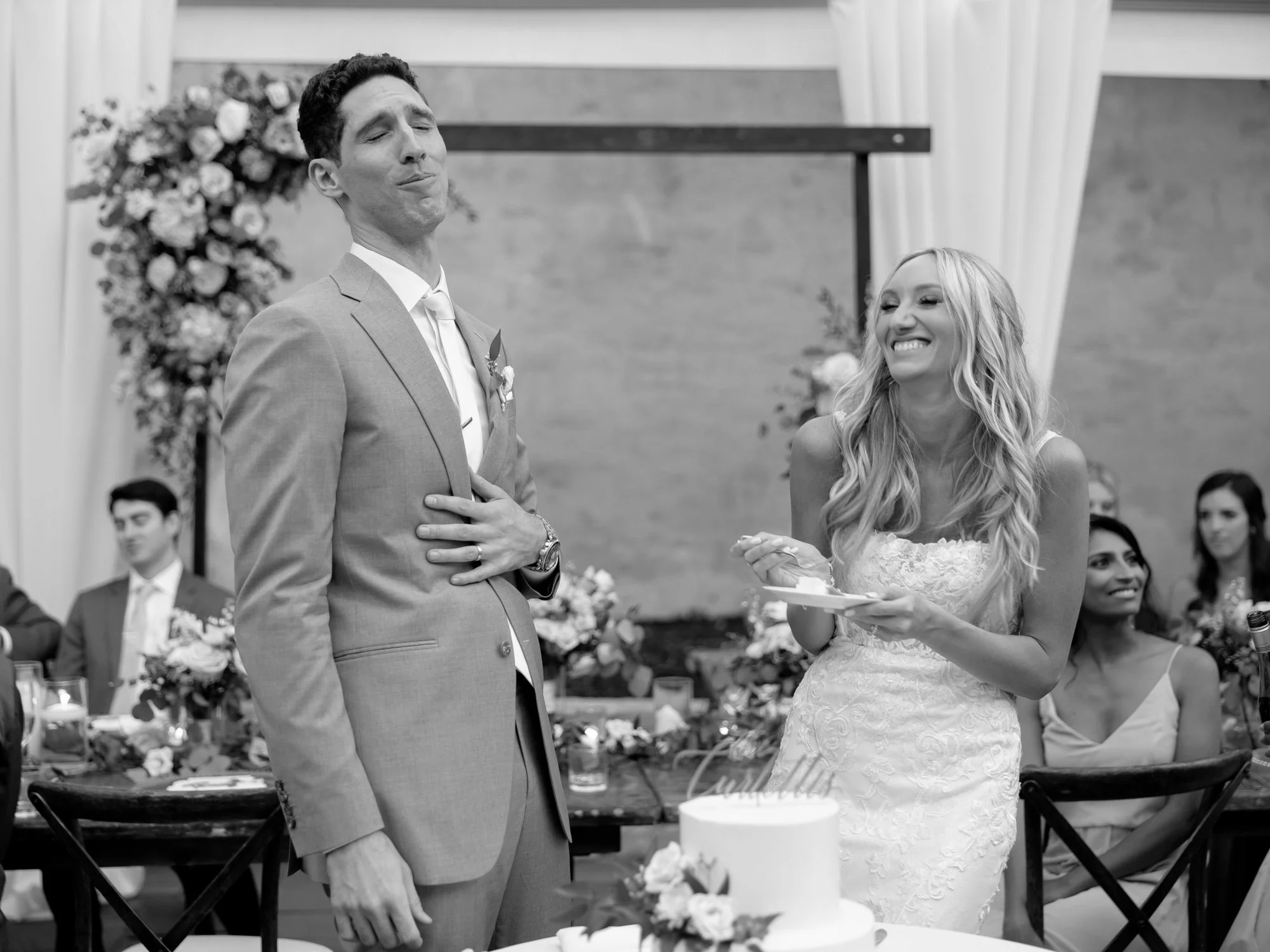 The groom makes a dramatic, playful reaction face at the cake cutting while the bride laughs beside him holding the plate, a candid black-and-white moment.
