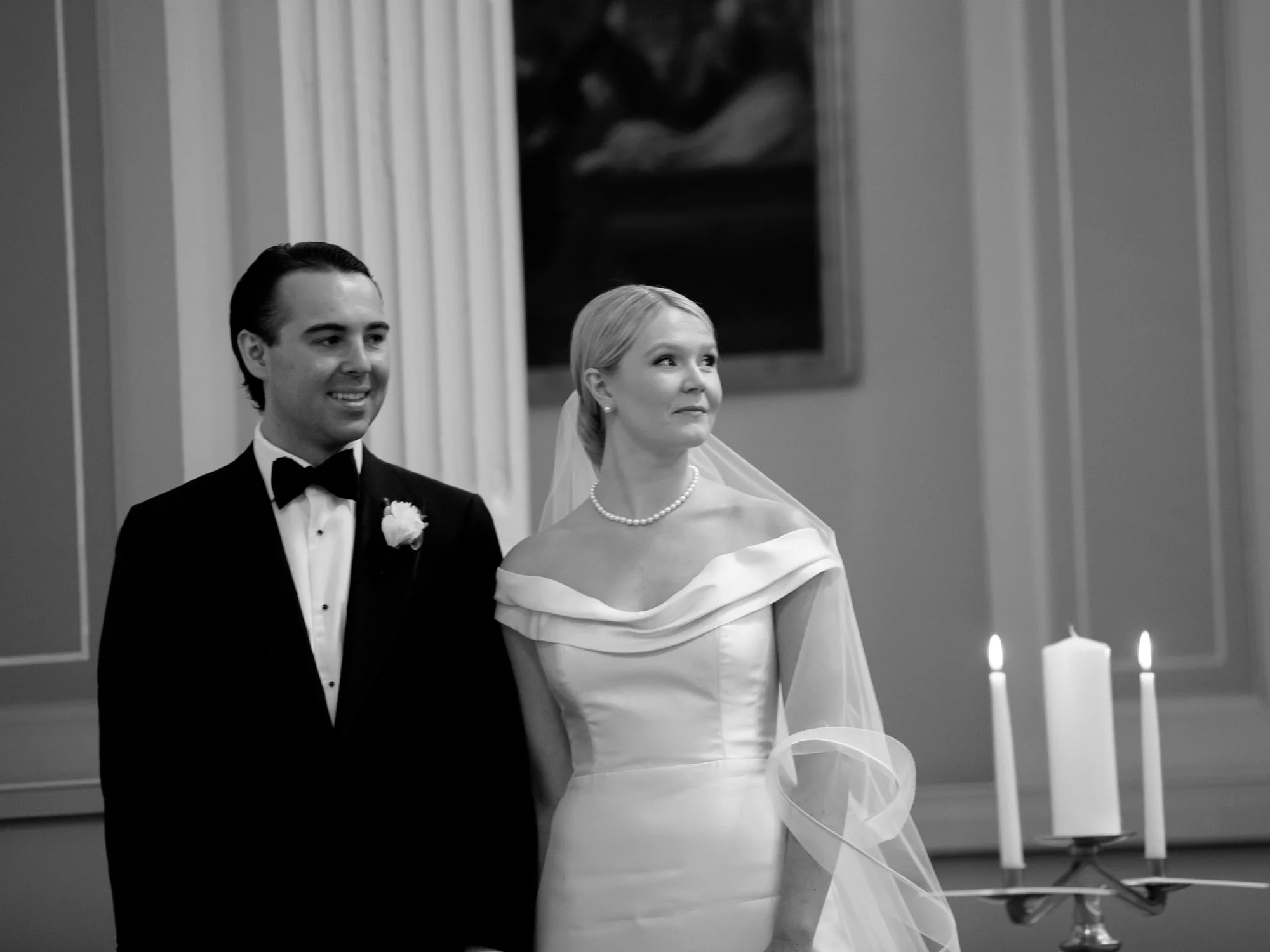 Bride and groom stand at the ceremony altar in black and white — pillar candles at their side, a classical oil painting rising behind them. Shot on film.