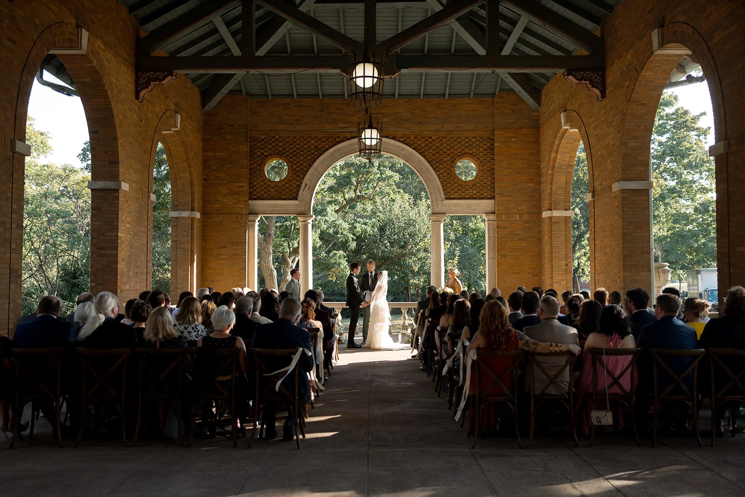 A sweeping ceremony wide shot from the back of the pavilion — the couple at the altar, every seat filled, the soaring brick arches of Columbus Park Refectory framing the moment. Chicago film wedding photographer.