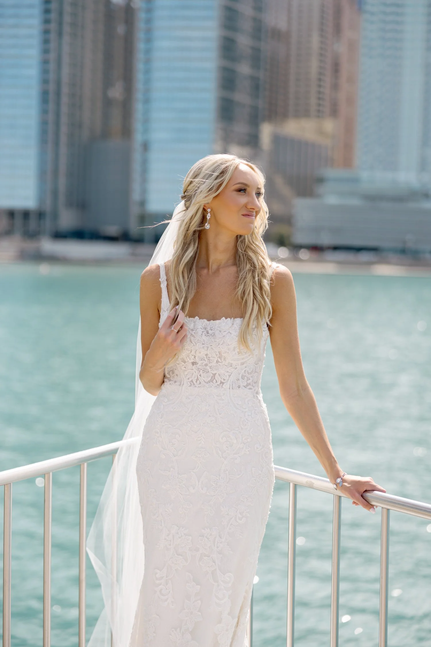The bride leans elegantly on a waterfront railing, her lace gown and loose veil catching the breeze as the gleaming Chicago skyline towers behind her.