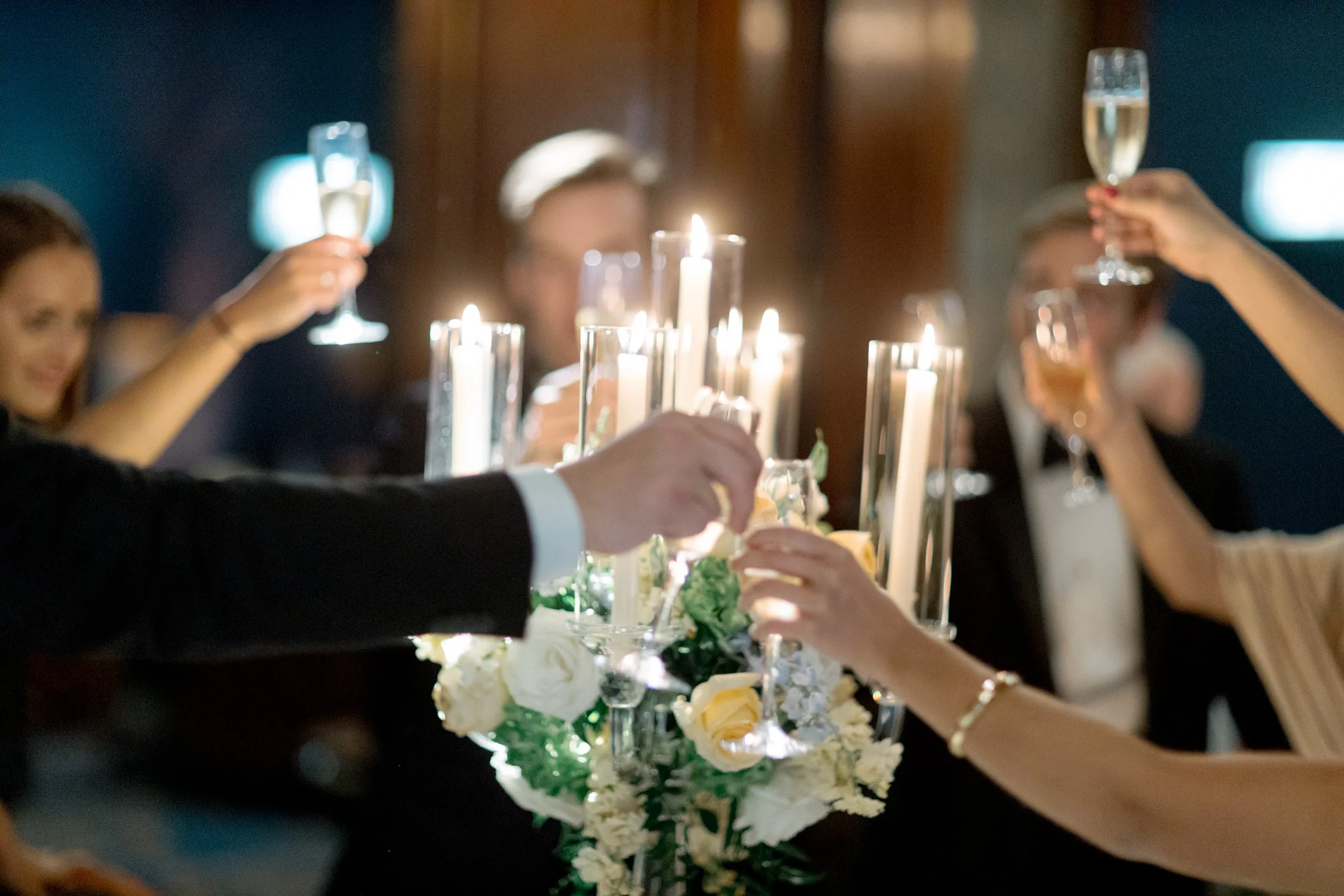 Wedding guests reach in to clink champagne flutes over a glowing floral centerpiece, taper candles and white roses just below the glasses mid-toast.