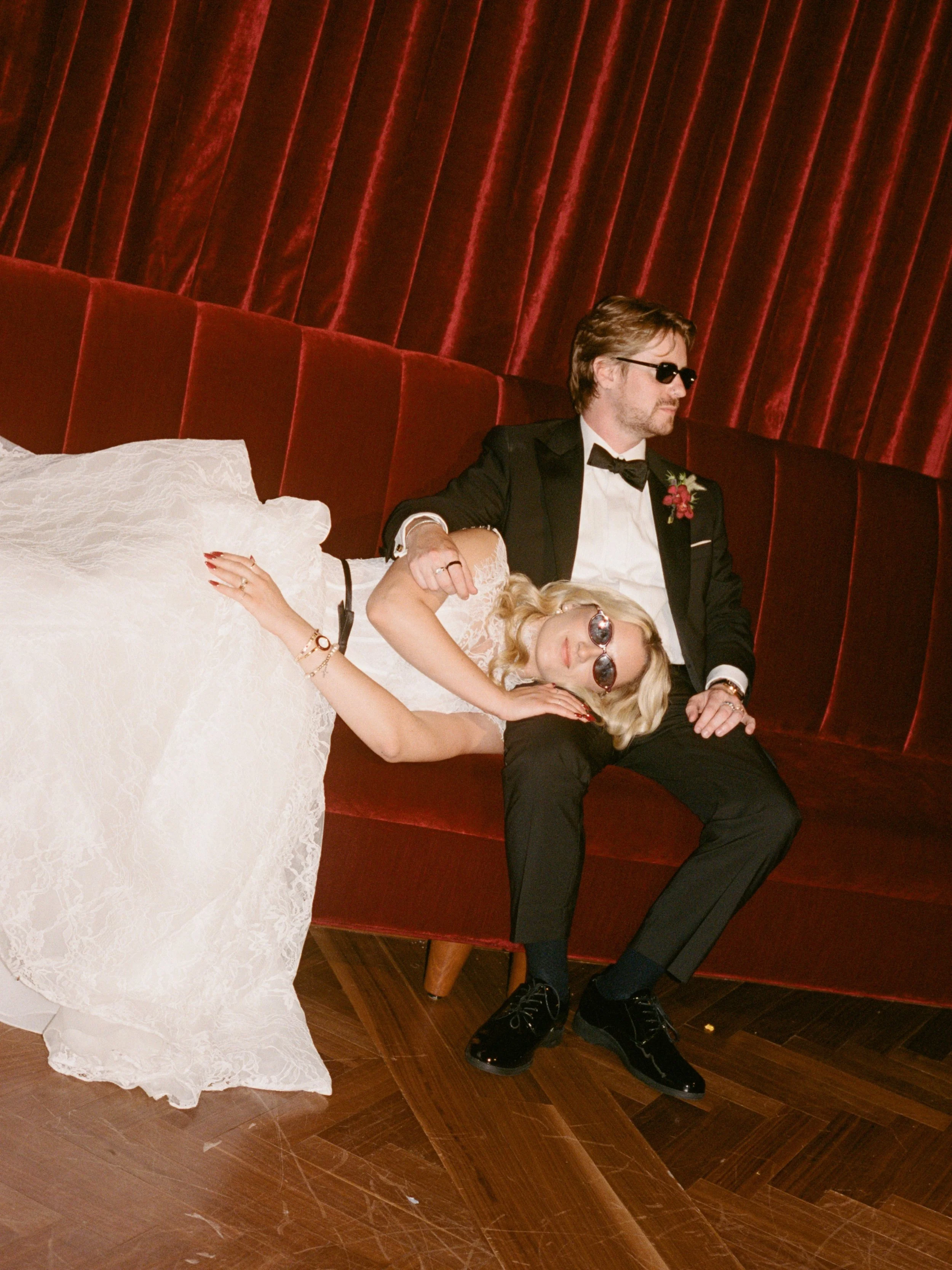 Bride reclines playfully across the groom's lap in sunglasses on the velvet couch in The Wellsley's red velvet lounge