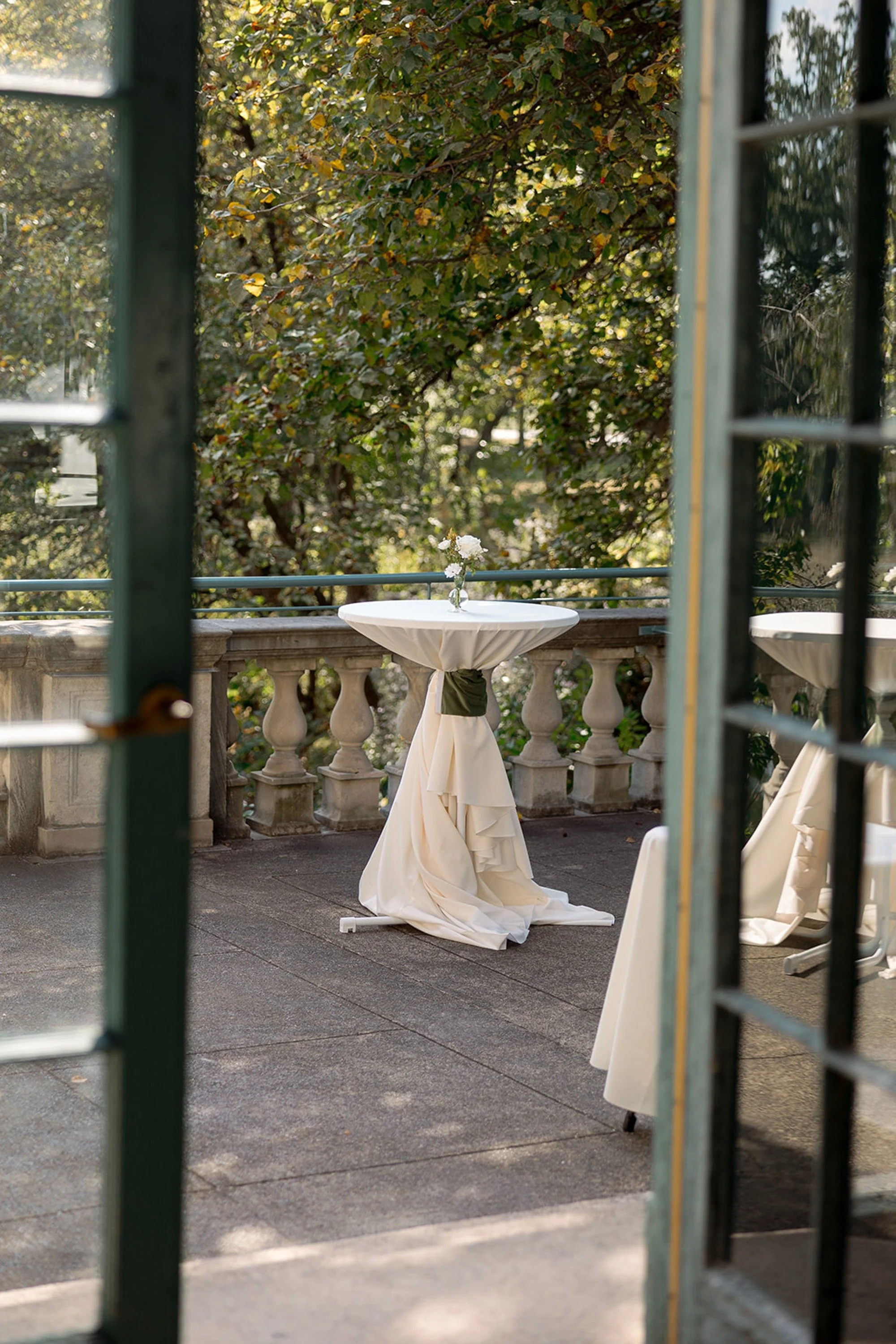 An ivory-draped cocktail table stands quietly on the stone terrace balcony of Columbus Park Refectory, framed by teal iron railings and a canopy of autumn foliage — the calm before the celebration. Chicago film wedding photographer.