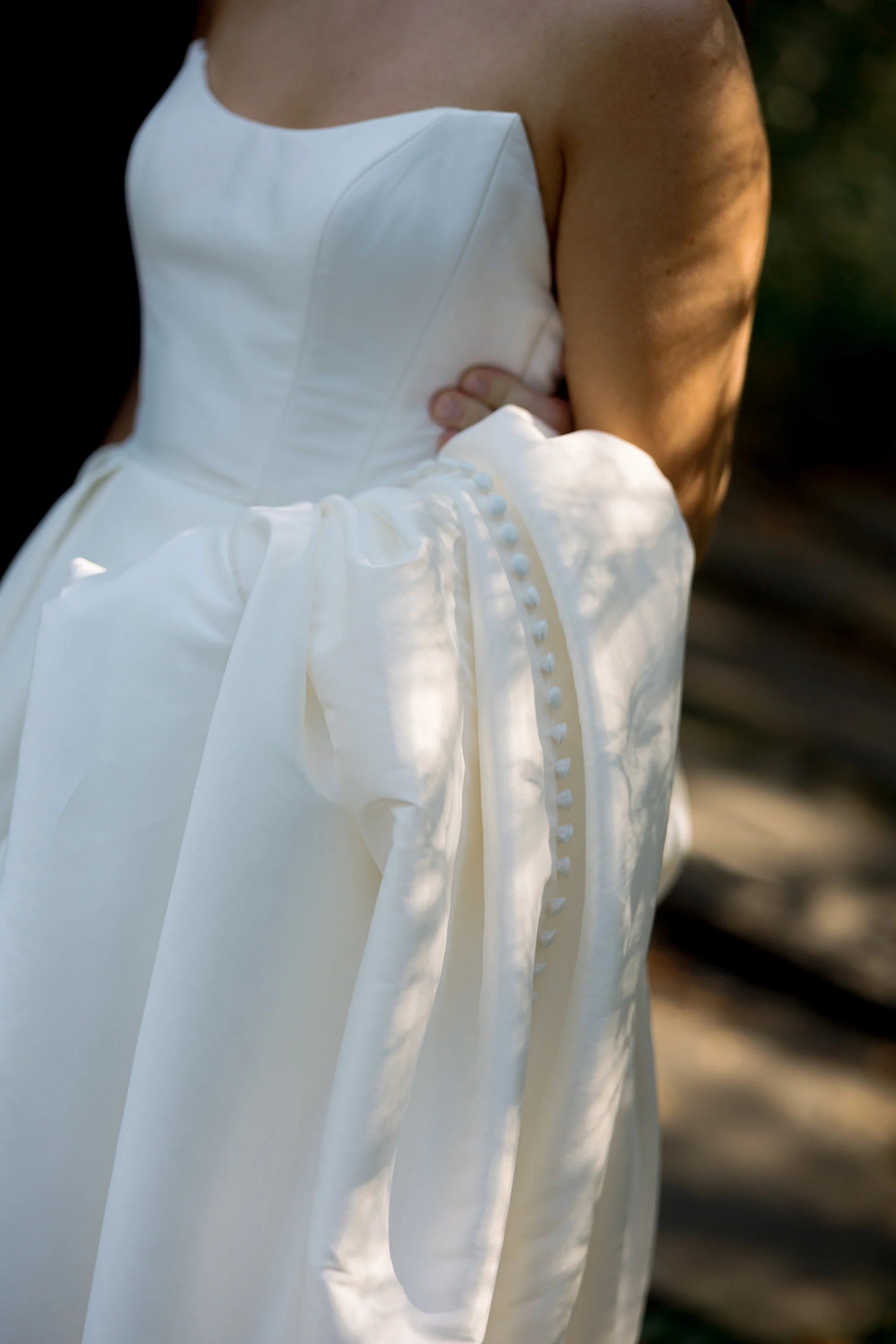 A dress detail shot in motion — the structured strapless bodice and luminous button-lined train of the bride's ballgown caught in a shaft of golden light at Columbus Park Refectory