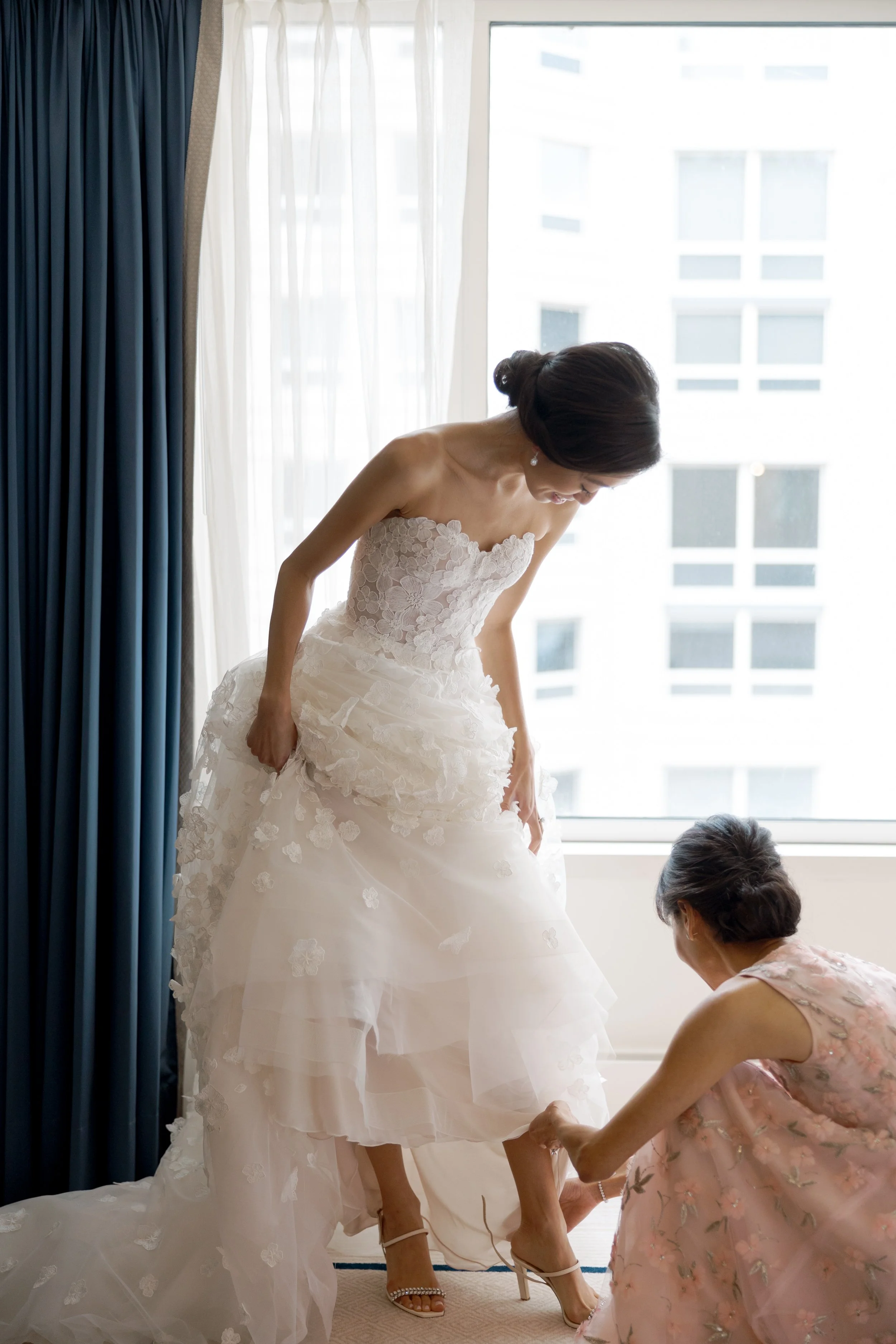 Brides mother kneels to fasten her wedding shoes by the hotel window at The Peninsula Chicago before the ceremony