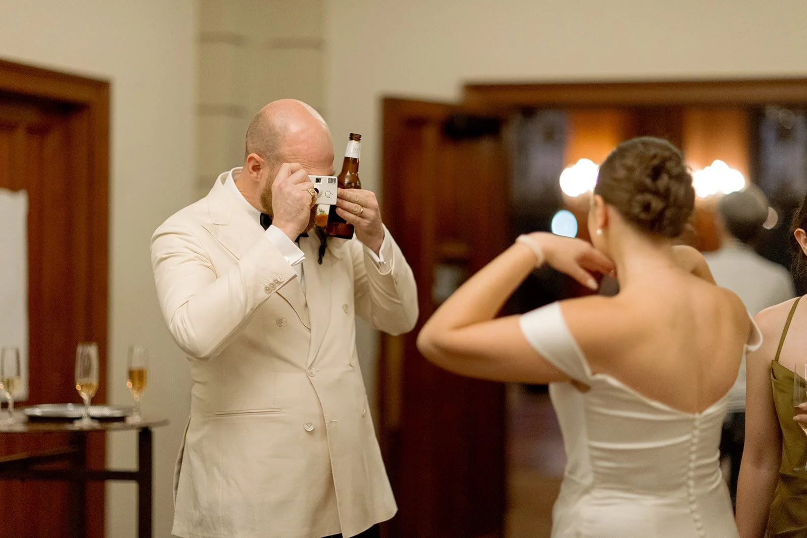 The groom, beer in hand, raises a 35mm camera to photograph his own bride during the reception — a meta, candid moment that captures who they are perfectly. Shot on film by a Chicago film wedding photographer.