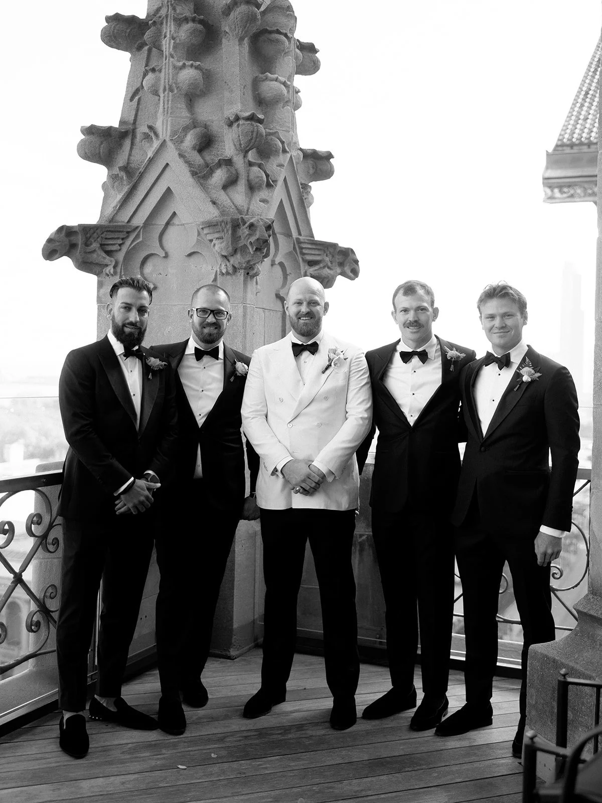 The groom and his groomsmen on the University Club Chicago rooftop terrace in black and white, a dramatic Gothic stone spire rising behind them.